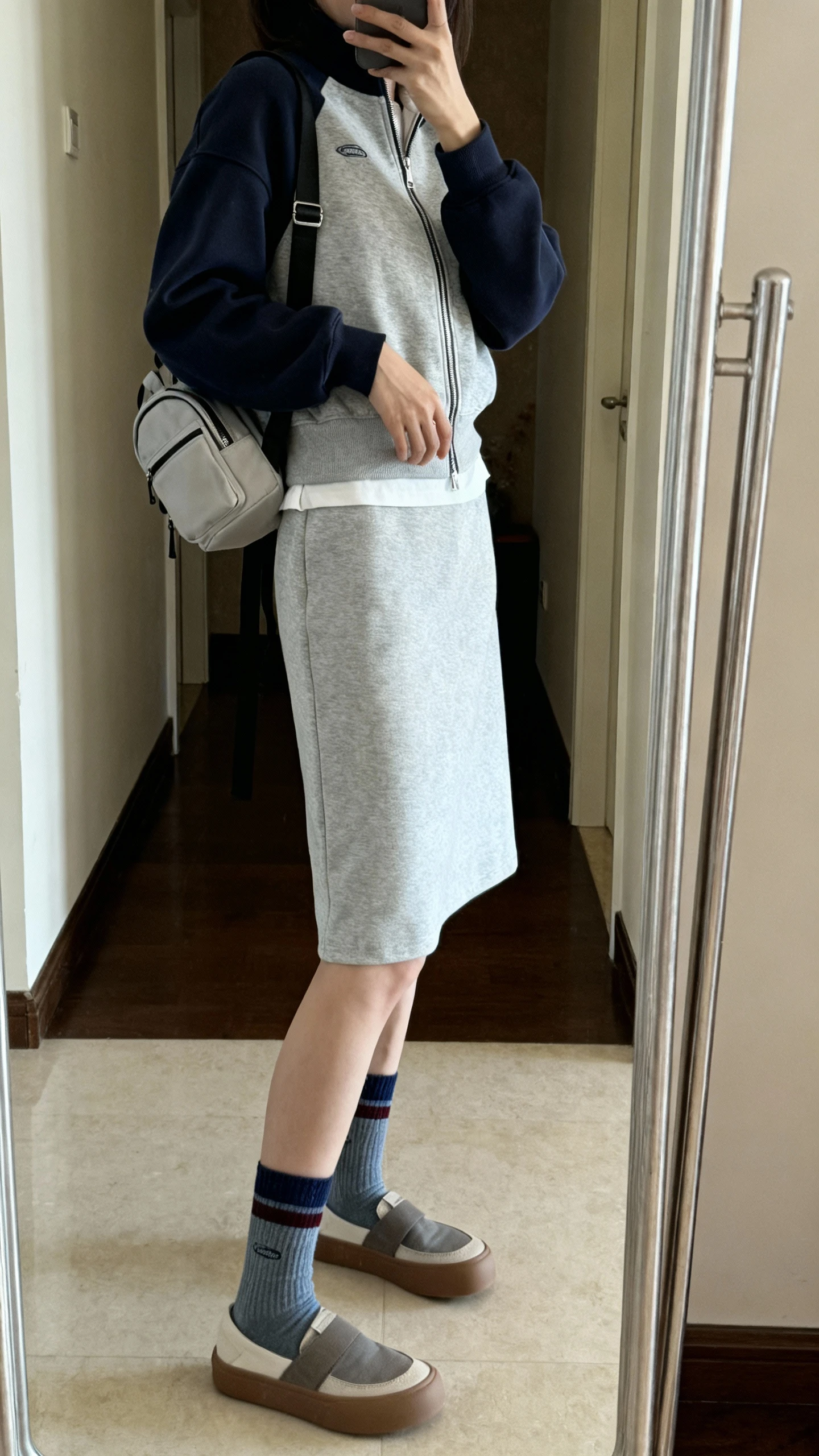 Casual iPhone mirror selfie of a woman in an athleisure dress layered with a zip fleece, sporty socks, slip-on sneakers, and a small backpack; face not visible, side angle, entryway full-length mirror, indoor natural light, iPhone photo quality.