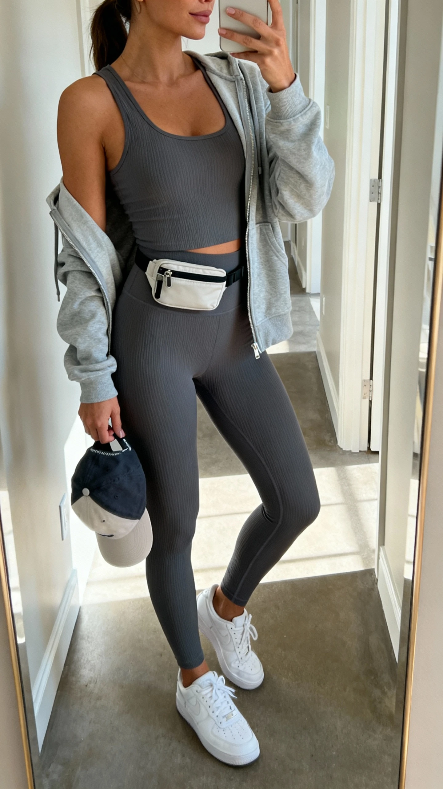 Casual iPhone mirror selfie of a woman wearing an athleisure minimalist set: matching ribbed tank and high-waist leggings in charcoal, lightweight zip-up hoodie, clean white sneakers, small crossbody or belt bag, baseball cap in hand; face not visible, side angle, hallway full-length mirror with natural daylight, iPhone photo quality.