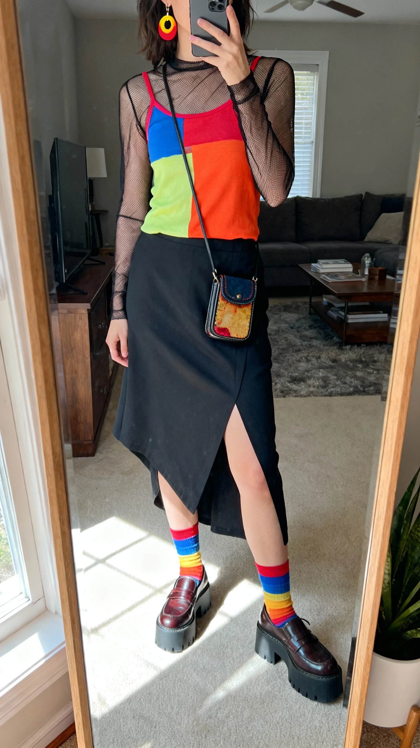 iPhone angled mirror selfie of a woman wearing artsy layers: sheer long-sleeve mesh top over a vibrant color-pop tank, asymmetric midi skirt, chunky platform loafers, color-block socks, bold statement earrings, and a quirky mini crossbody, face not visible, living room full-length mirror with natural daylight, iPhone photo quality.