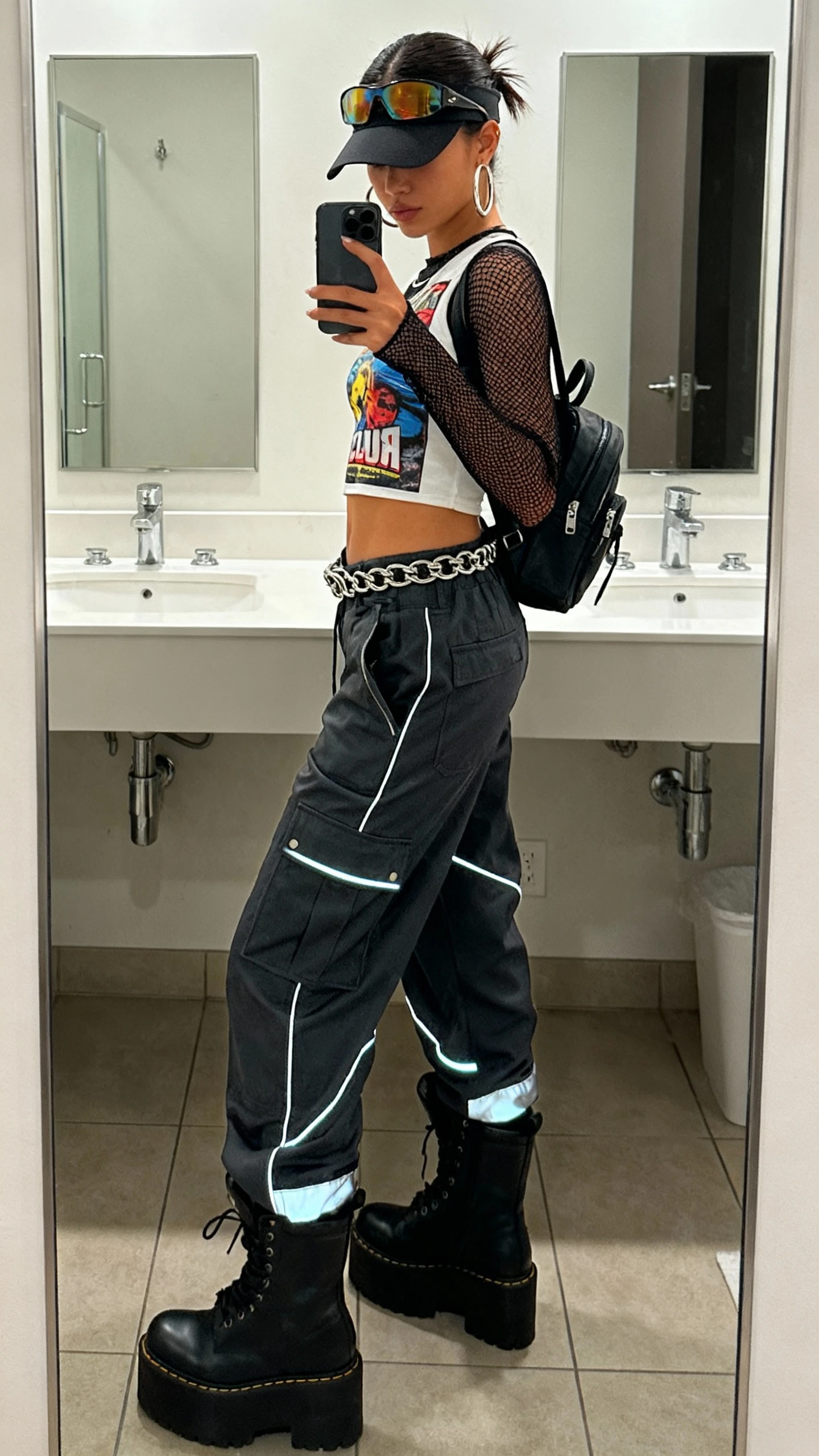 iPhone back/side-angle mirror selfie of a woman wearing a cyber club kid streetwear remix: mesh long-sleeve top under a cropped graphic tank, low-slung cargo pants with reflective piping, chunky platform boots, chain belt, mini backpack, tinted visor sunglasses pushed to head, and chunky hoop earrings, face not visible, bathroom mirror, indoor lighting, iPhone photo quality