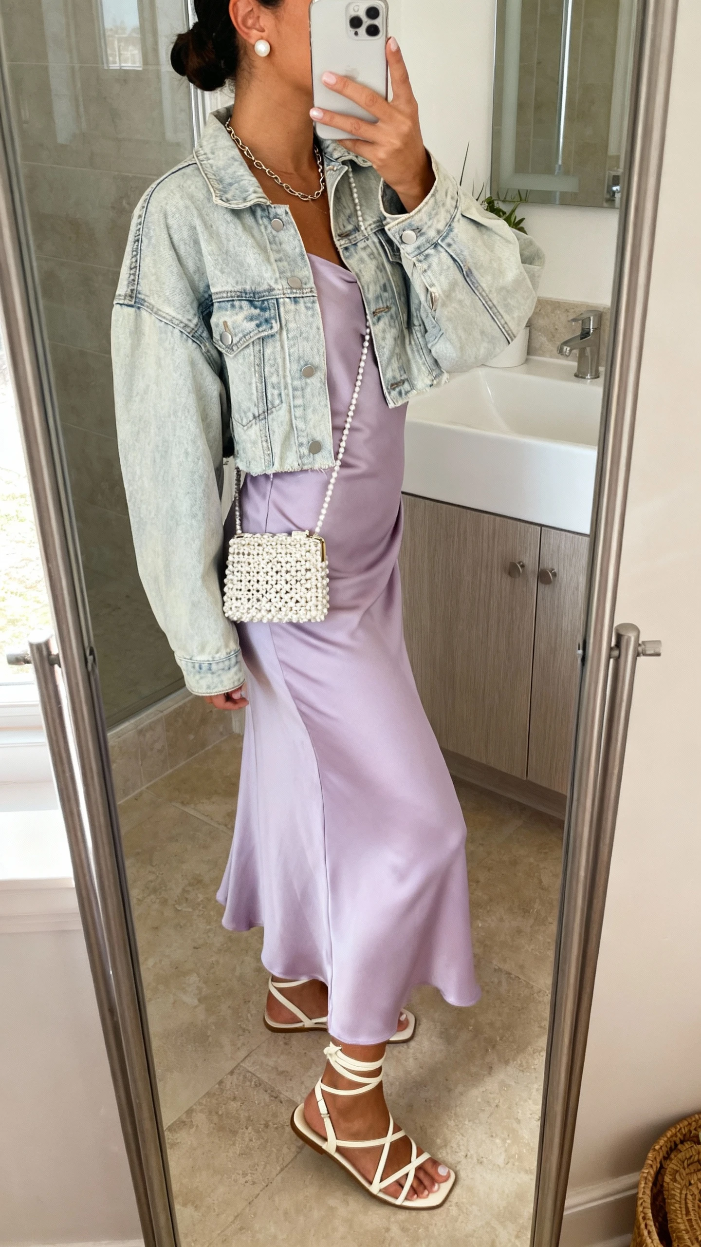 iPhone back/side mirror selfie of a woman in a bias-cut lavender midi slip dress with a light-wash cropped boxy denim jacket, strappy sandals in cream, a beaded mini bag, pearl studs, and a thin chain necklace, face not visible, bathroom mirror, soft morning light, iPhone photo quality.