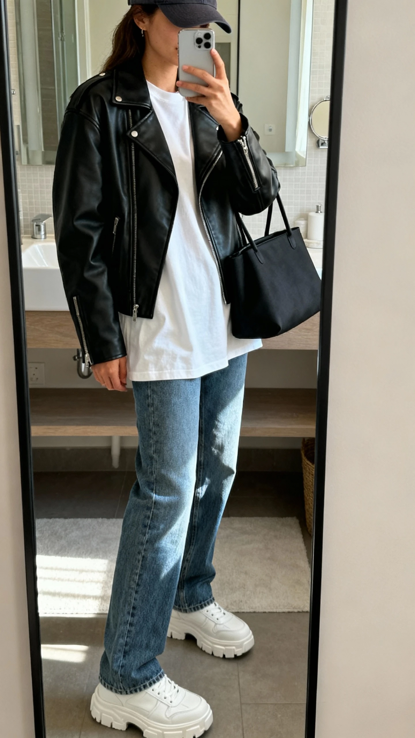 iPhone back/side mirror selfie of a woman wearing mom jeans with a boxy white tee, black leather moto jacket, chunky white sneakers, baseball cap, and a simple black tote, face not visible, bathroom full-length mirror, indoor morning light, iPhone photo quality.