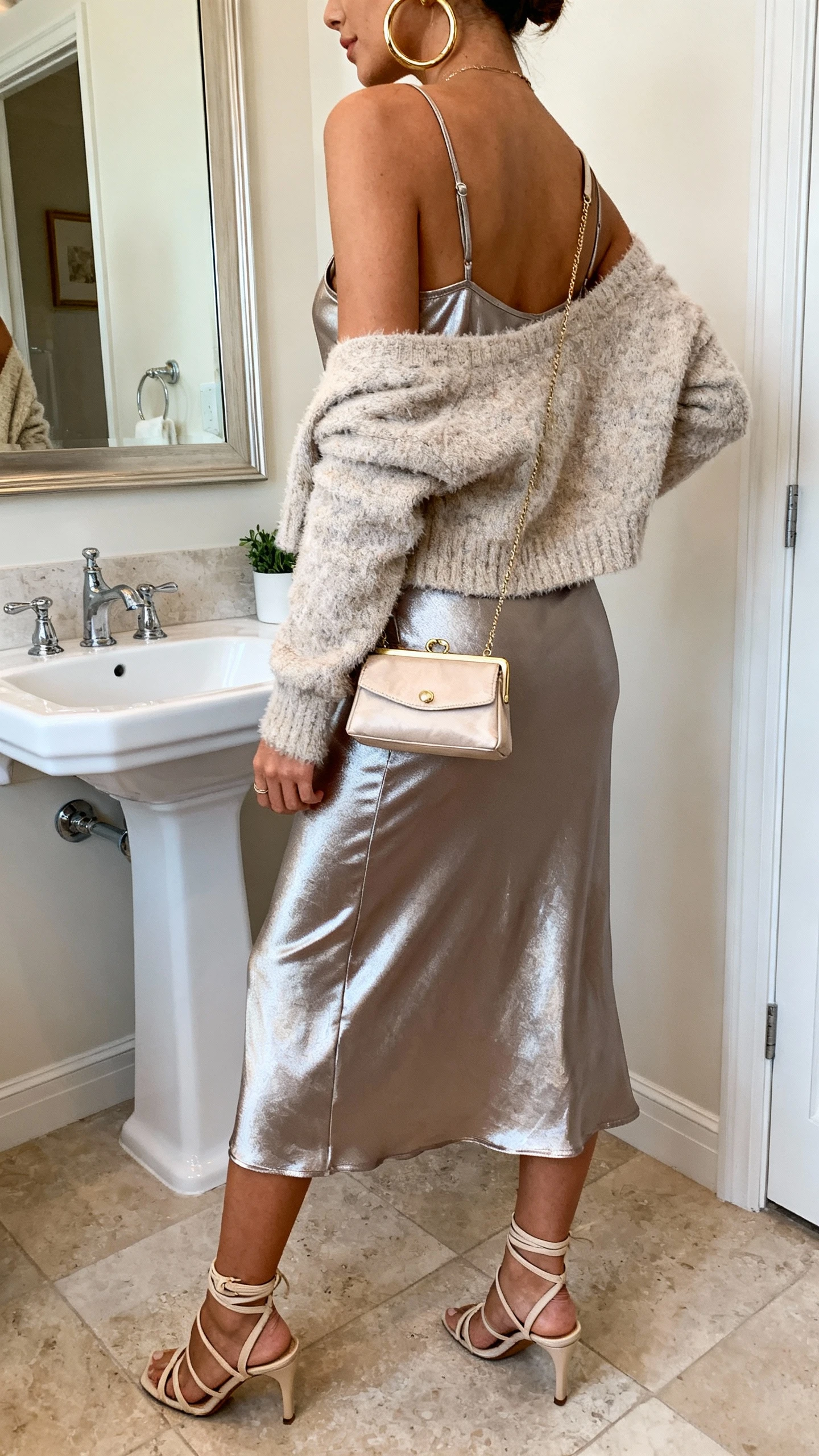 iPhone back/side view of a woman wearing a metallic slip dress layered with a fuzzy cropped cardigan, strappy heels, mini baguette bag, and chunky hoop earrings, face not visible, bathroom full-length mirror, soft indoor lighting, iPhone photo quality