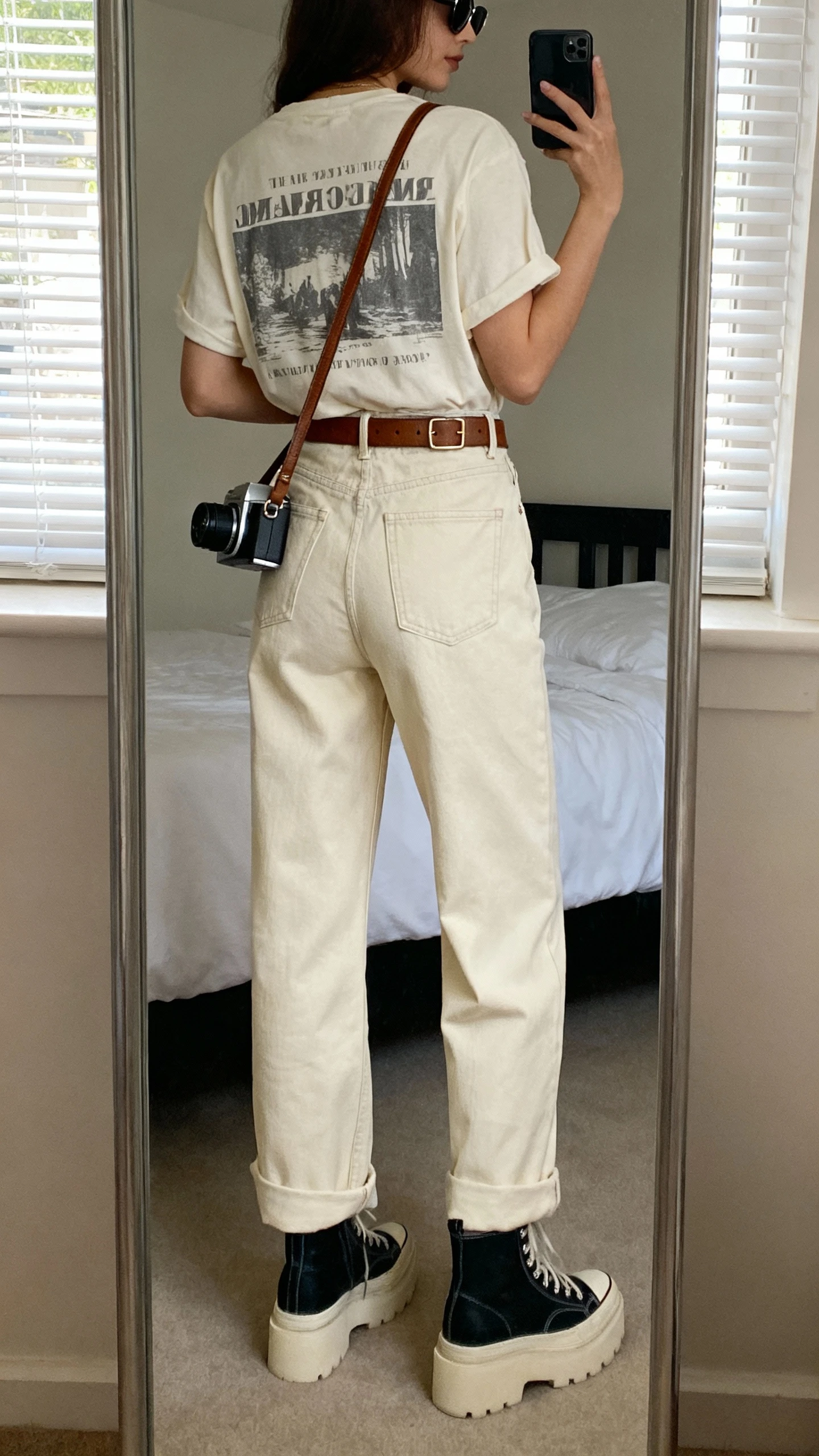 iPhone back-view mirror selfie of a woman in a soft graphic tee (vintage band/minimal typography/artsy print) tucked into high-rise wide-leg jeans (medium or ecru wash), chunky platform sneakers or ankle boots, a leather belt with subtle hardware, a crossbody camera bag or mini shoulder bag, and sleek sunglasses; face not visible, bedroom mirror, window light, casual iPhone photo.