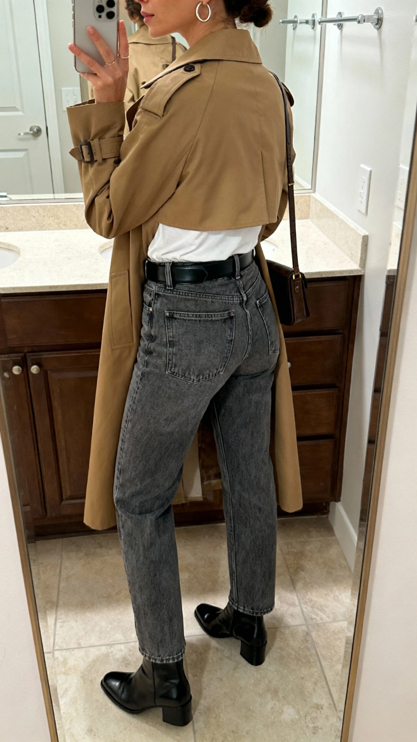 iPhone back-view mirror selfie of a woman wearing elevated denim and a trench: classic camel trench coat, high-rise mid-wash straight jeans, white tee, black leather belt, polished ankle boots, simple hoop earrings, crossbody bag; face not visible, bathroom mirror, soft indoor lighting, iPhone photo quality.