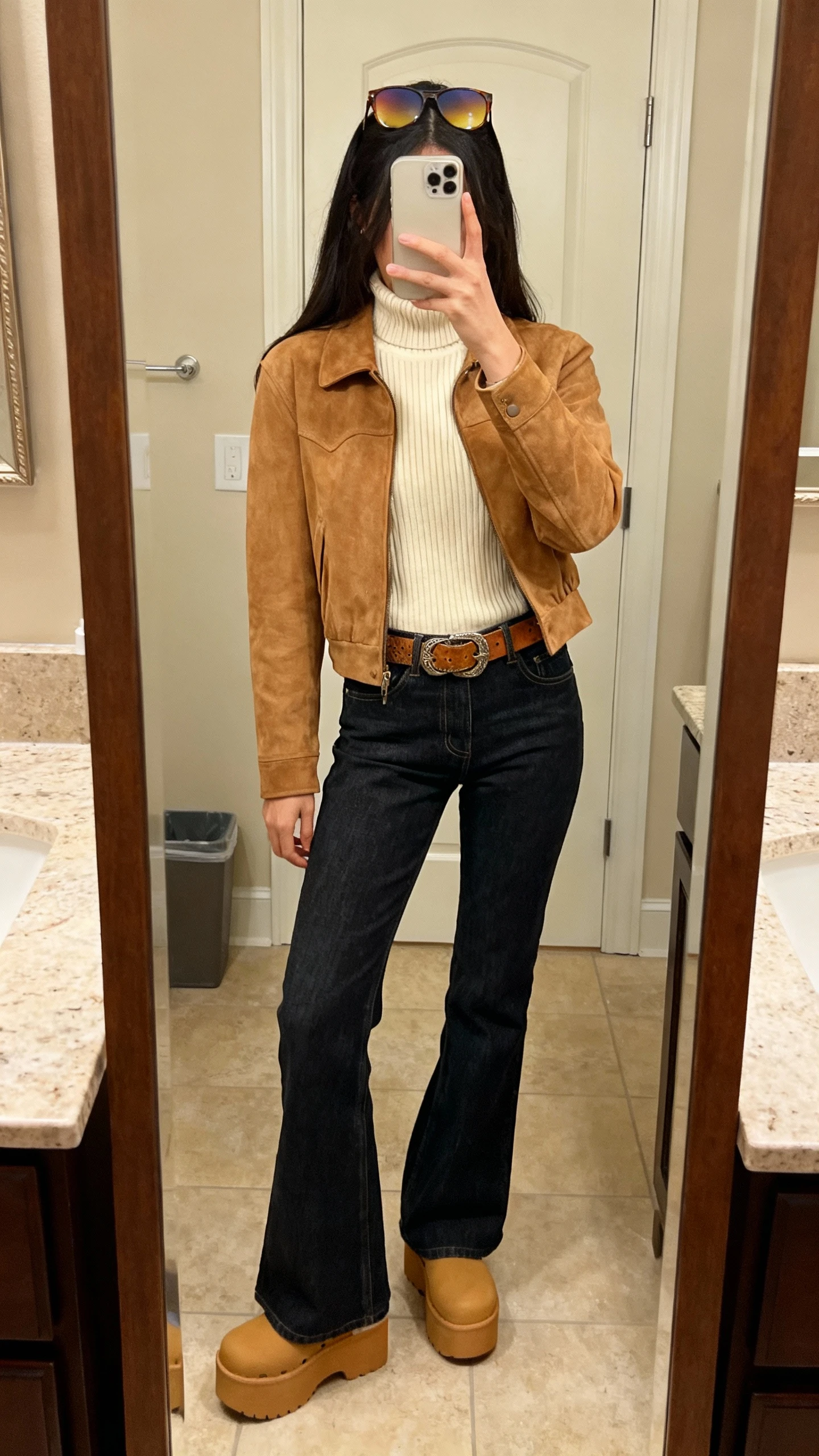 iPhone back-view mirror selfie of a woman wearing dark-wash flared denim, a camel suede jacket over a ribbed cream turtleneck, tan platform clogs, oversized gradient sunglasses pushed up on head, and a tooled leather belt, face not visible, bathroom full-length mirror, warm indoor lighting, iPhone photo quality.