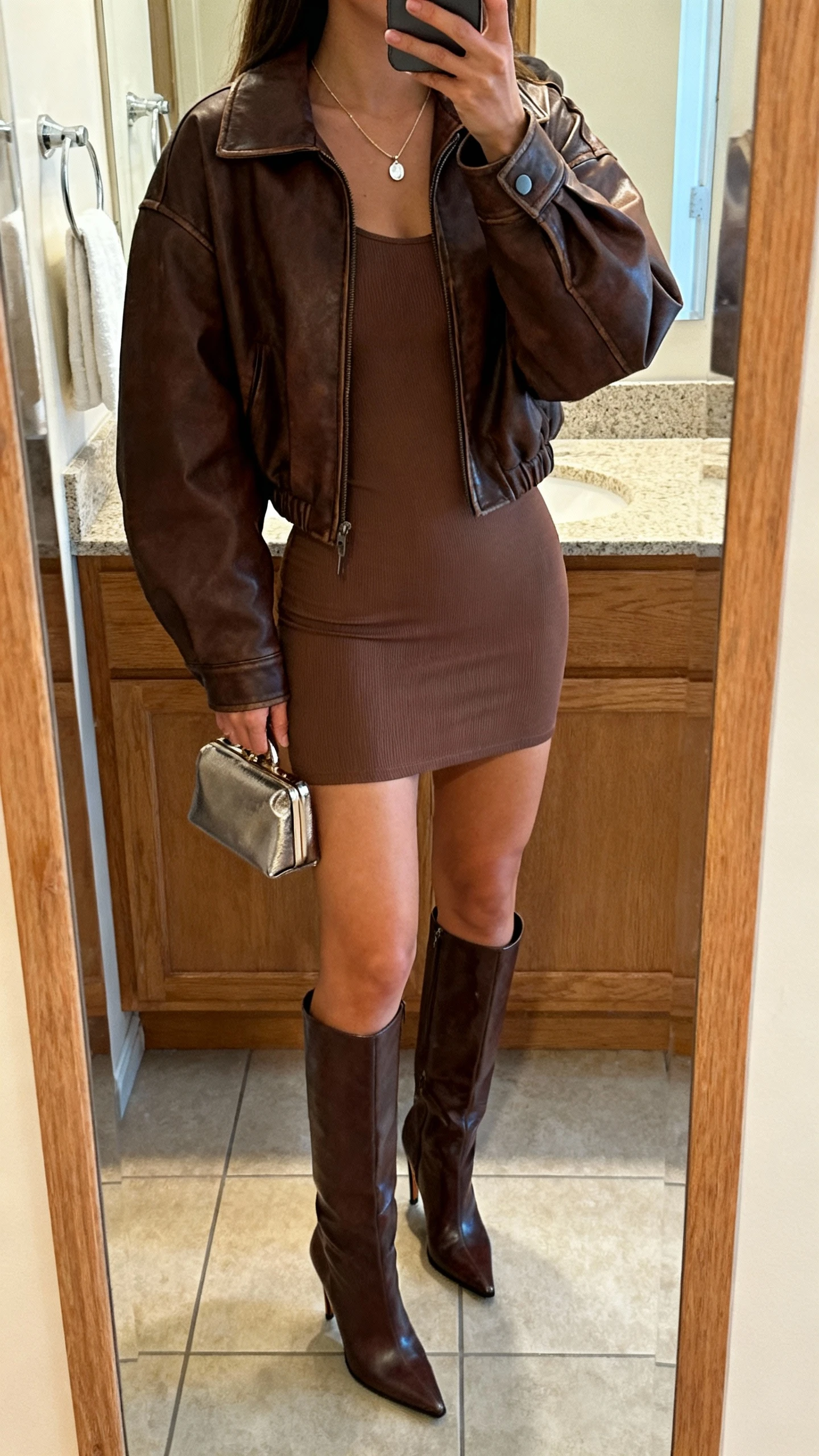 iPhone back-view mirror selfie of a woman wearing a chocolate leather jacket over a fitted bodycon mini dress and knee-high heeled boots, small metallic clutch, delicate pendant necklace, face not visible, bathroom mirror, warm indoor lighting, iPhone photo quality.