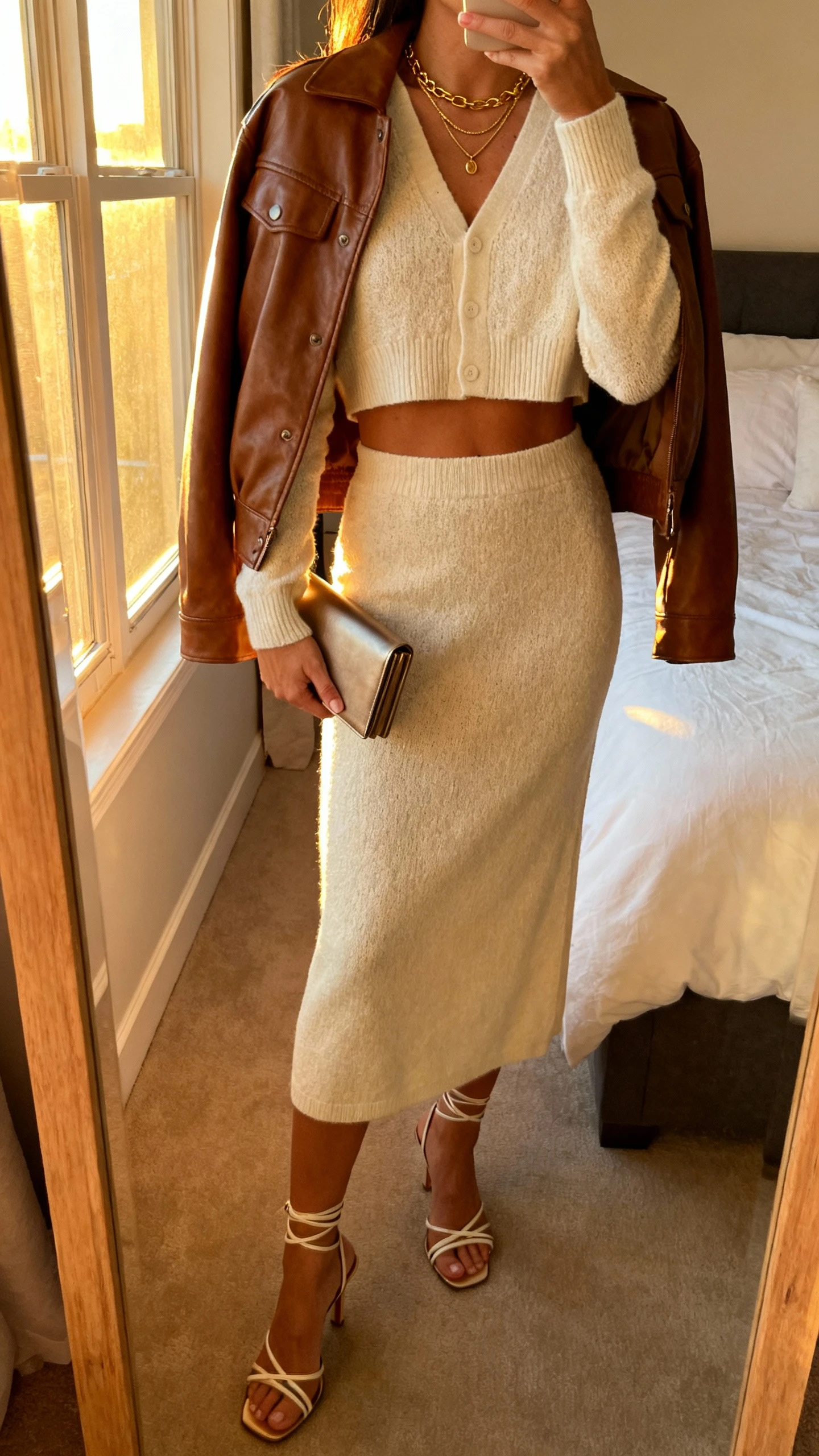 iPhone back-view mirror selfie of a woman wearing a brown leather jacket draped over a cream knit skirt set (cropped cardigan top and midi skirt), strappy heeled sandals, delicate layered gold necklace, slim clutch, face not visible, golden hour window light in bedroom, iPhone photo quality