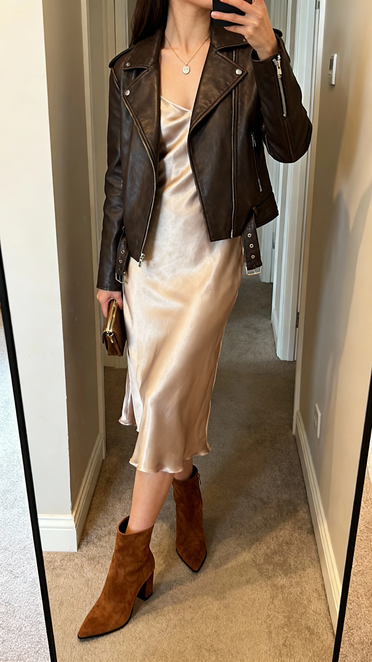 iPhone back-view mirror selfie of a woman wearing suede heeled ankle boots, slinky satin slip dress, tailored leather jacket, delicate pendant necklace, and a small clutch in hand, face not visible, hallway full-length mirror with warm indoor lighting, iPhone photo quality.