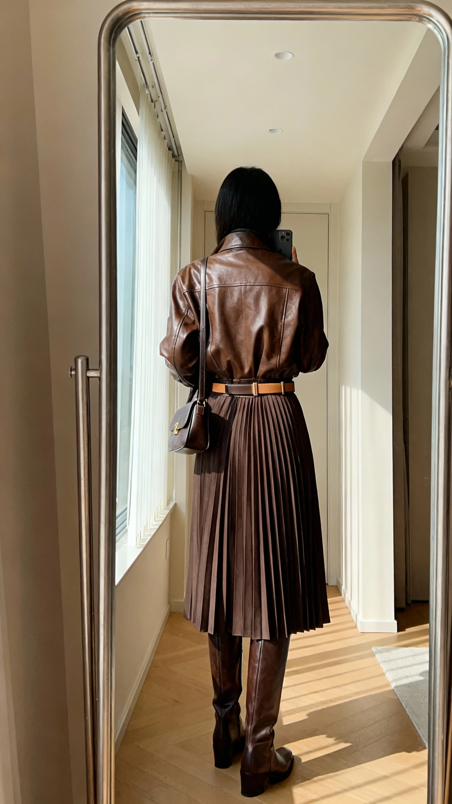 iPhone back-view mirror selfie of a woman wearing a brown leather jacket over a pleated midi skirt, tall knee-high boots, slim belt, structured shoulder bag, face not visible, full-length hallway mirror, morning window light, iPhone photo quality
