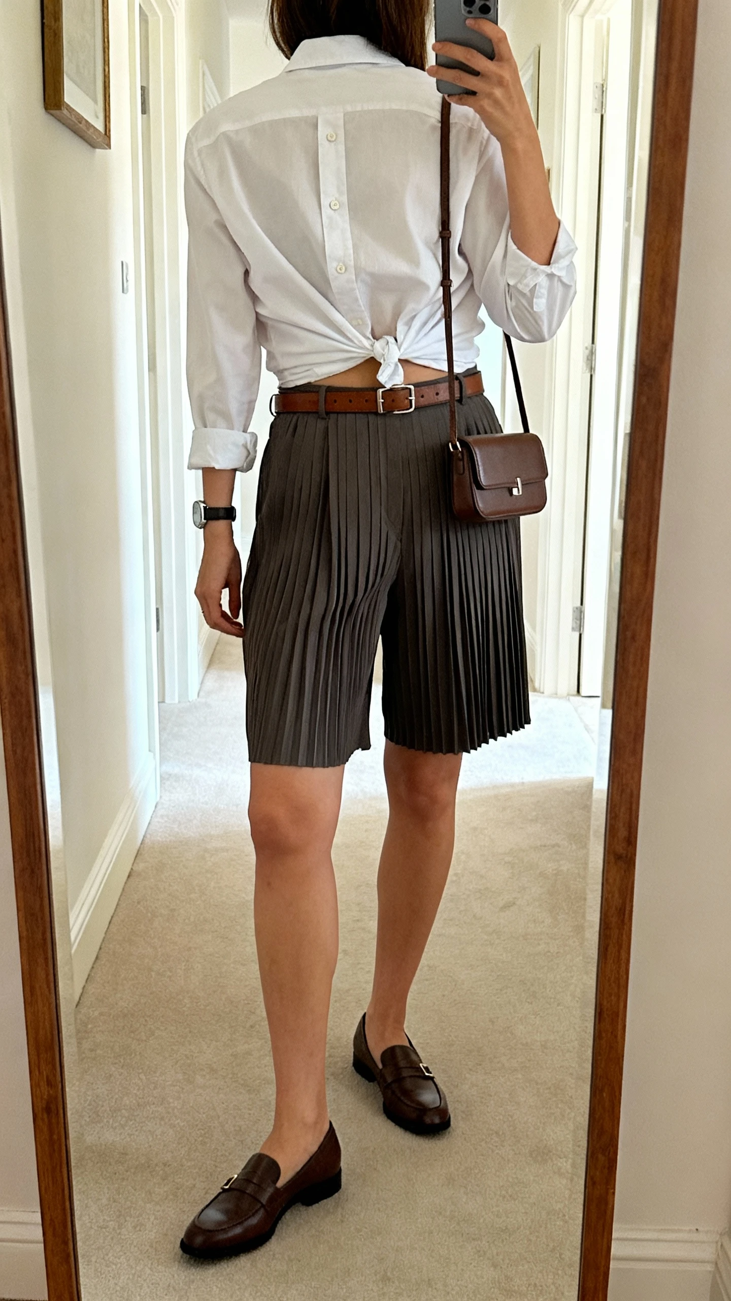iPhone back-view mirror selfie of a woman wearing tailored pleated shorts and a crisp button-up shirt half-tucked, leather belt, loafers, structured mini crossbody, minimalist watch, face not visible, full-length mirror in hallway, indoor daylight, iPhone photo quality.