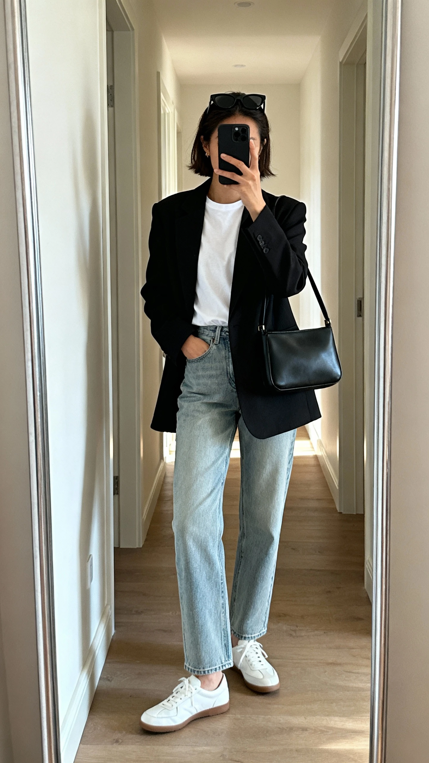 iPhone back-view mirror selfie of a woman wearing straight-leg light-wash jeans, a tailored black blazer over a white tee, white leather sneakers, black sunglasses perched on head, and a structured black shoulder bag, face not visible, hallway full-length mirror, indoor daylight, iPhone photo quality