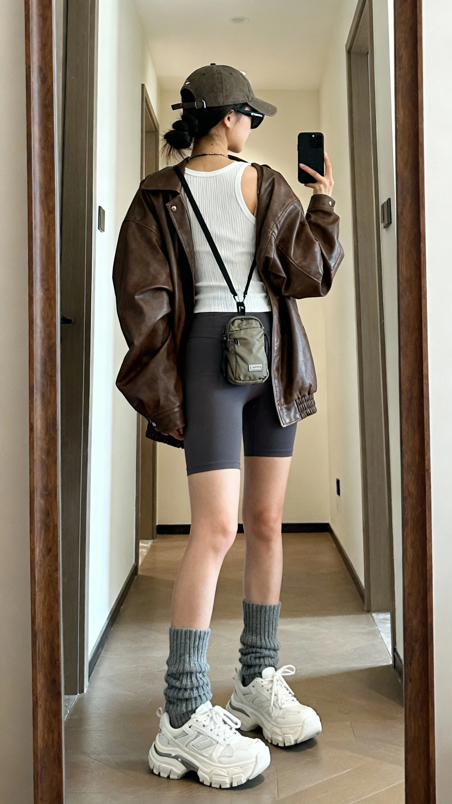 iPhone back view mirror selfie of a woman wearing a brown leather oversized jacket over a white ribbed tank, charcoal biker shorts, chunky gray crew socks with white dad sneakers, a baseball cap, crossbody nylon mini bag, and slim black sunglasses, face not visible, full-length mirror in hallway, natural daylight, iPhone photo quality