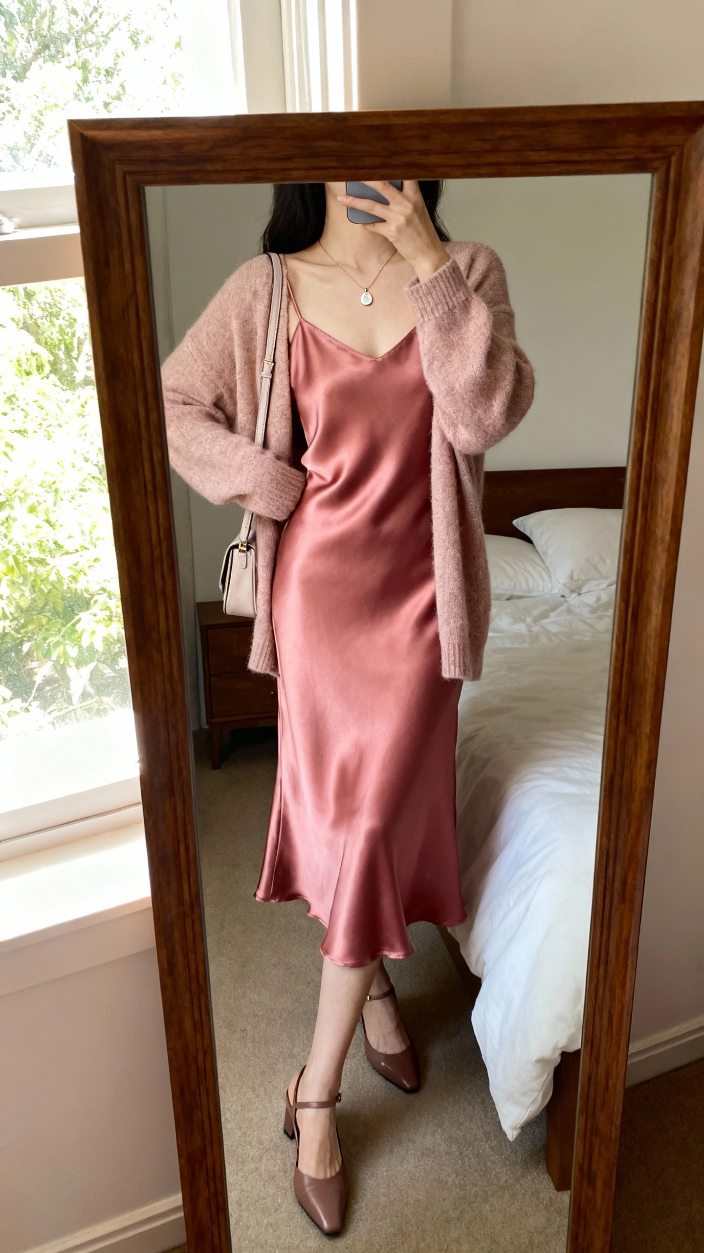 iPhone back-view mirror selfie of a woman wearing a rosy satin slip dress under a soft cardigan, delicate pendant necklace, slingback heels, and a small shoulder bag, face not visible, bedroom mirror with window light, casual iPhone photo quality.