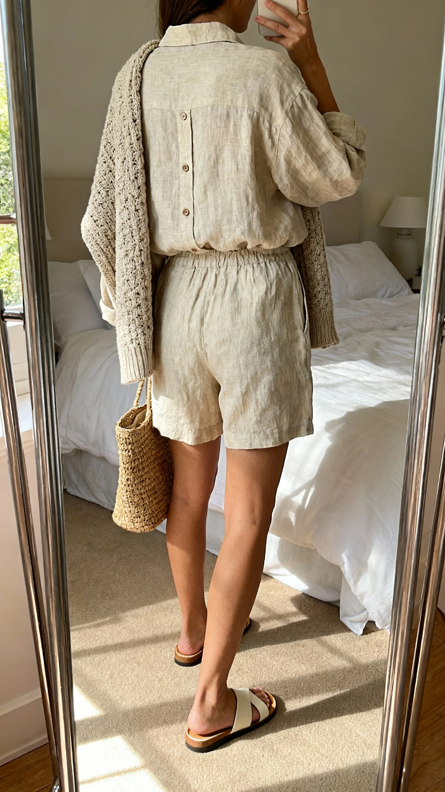 iPhone back-view mirror selfie of a woman wearing a matching linen shorts set (button-up shirt and relaxed shorts) layered with a chunky cardigan draped over shoulders, slide sandals, and a woven tote, face not visible, bedroom mirror with soft afternoon light, iPhone photo quality.