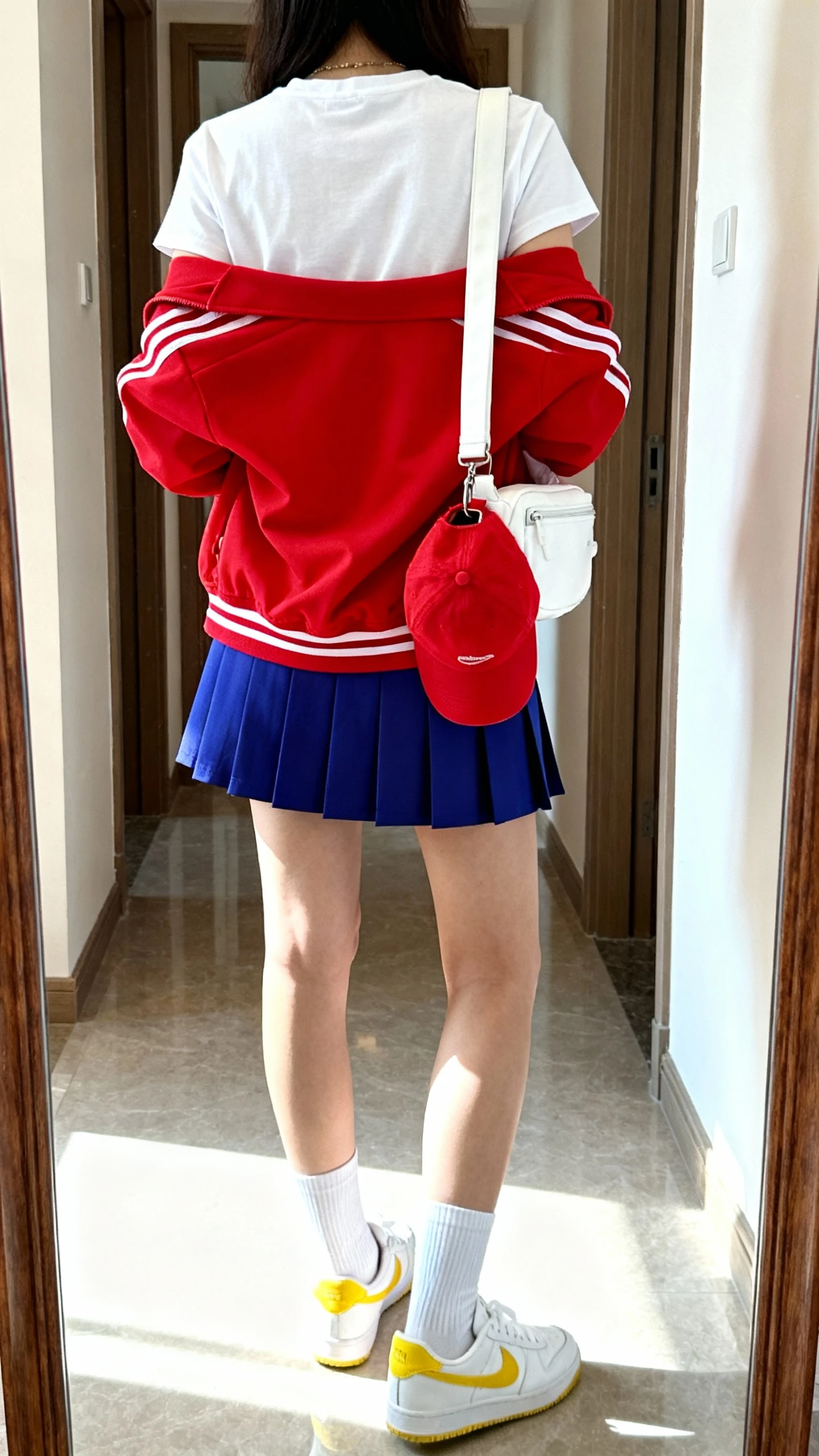 iPhone back view mirror shot of a woman in retro-sport pop: crisp white tee, primary-red track jacket, cobalt blue pleated mini skirt, white crew socks, white sneakers with yellow accents, red cap clipped to a white sling bag; face not visible, hallway mirror, bright daylight, iPhone photo quality.