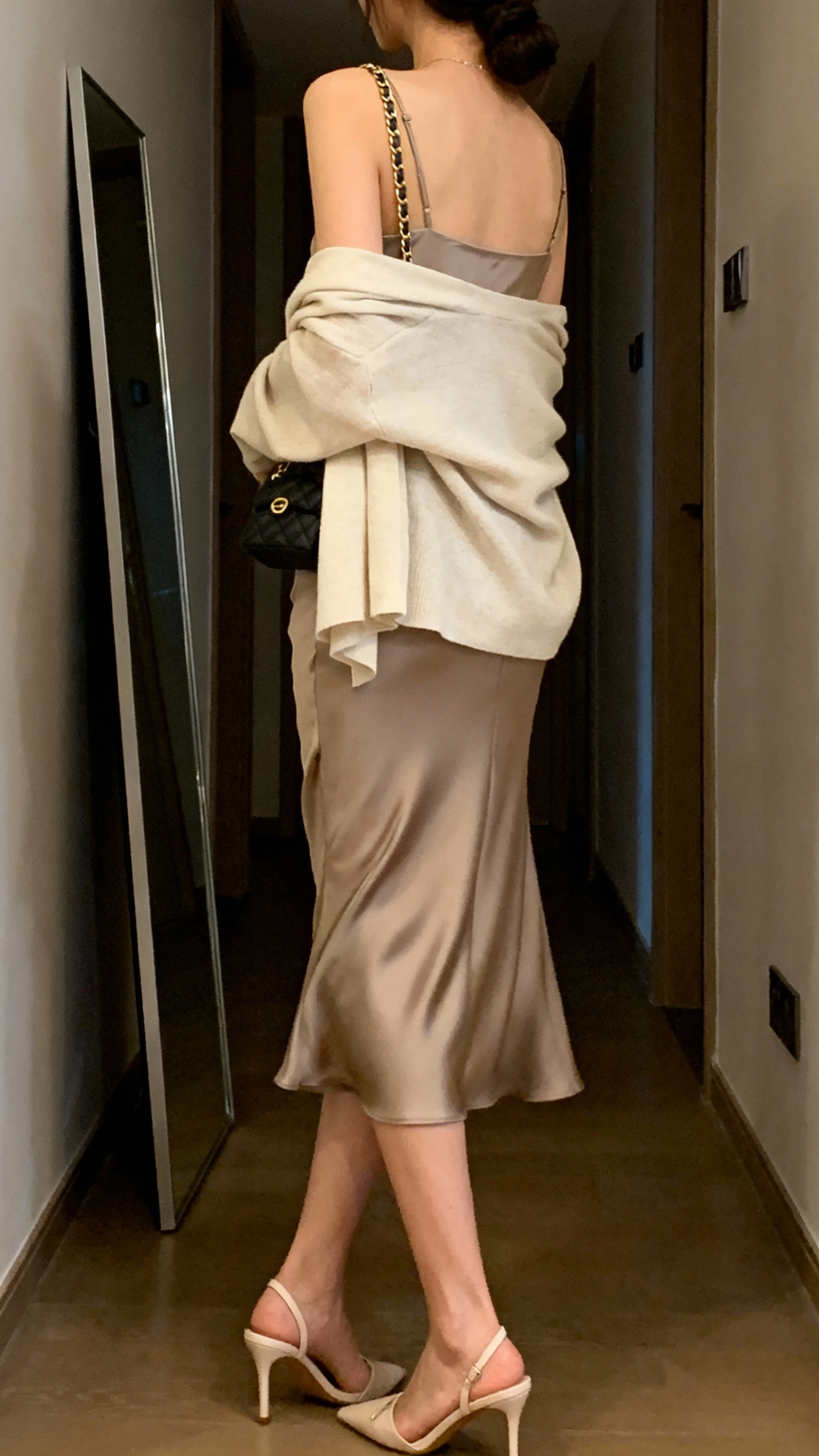 iPhone back-view mirror shot of a woman wearing a bias-cut slip dress with a light cardigan draped over shoulders, delicate heels, and a chain-strap shoulder bag; face not visible, hallway full-length mirror, soft evening indoor light, iPhone photo quality.
