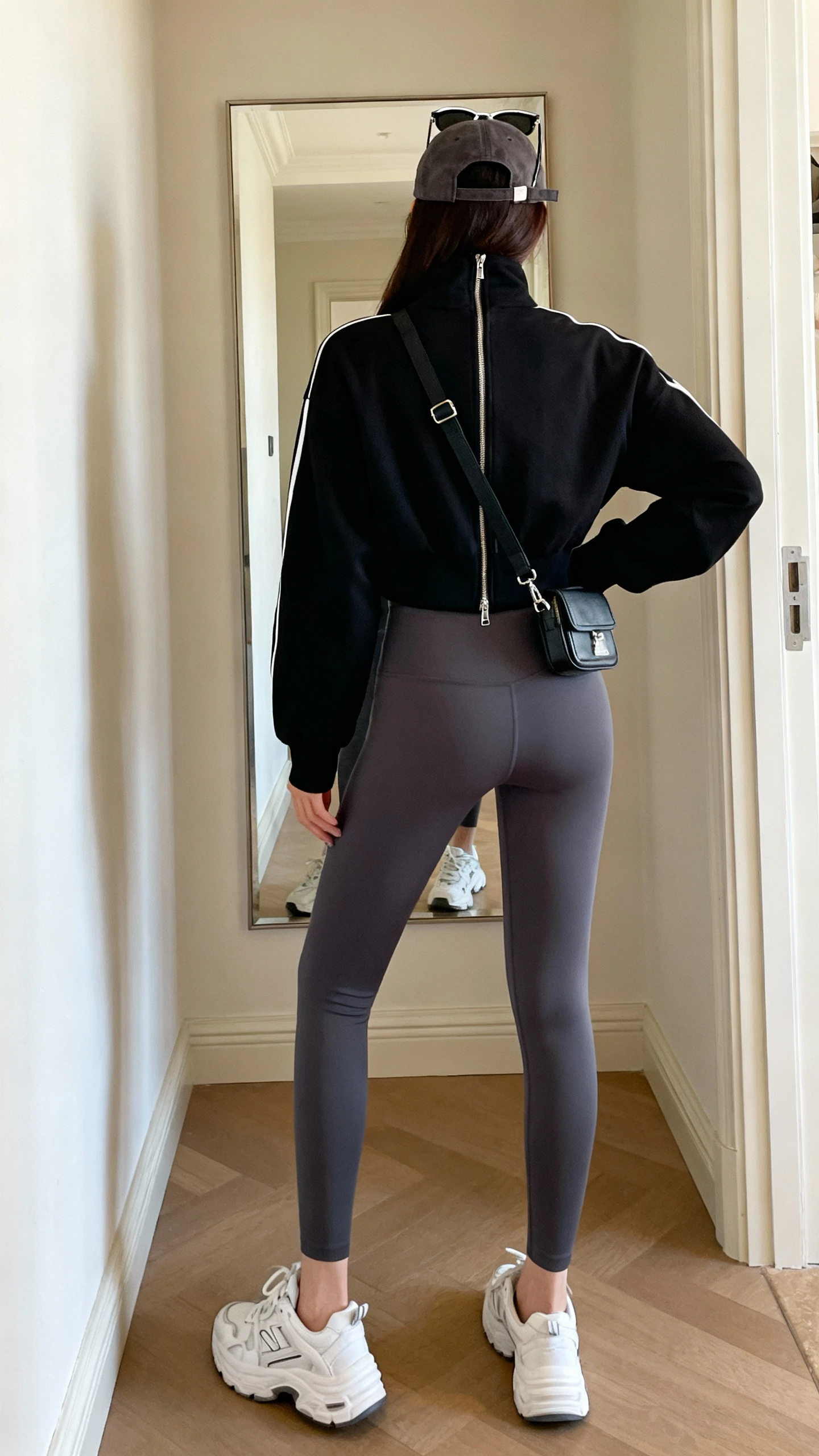 iPhone back view of a woman in a black zip-up track jacket and charcoal high-waist leggings, white dad sneakers, baseball cap, mini belt bag worn crossbody, oversized sunglasses pushed to head, face not visible, hallway mirror after errands, indoor daylight, casual iPhone photo.