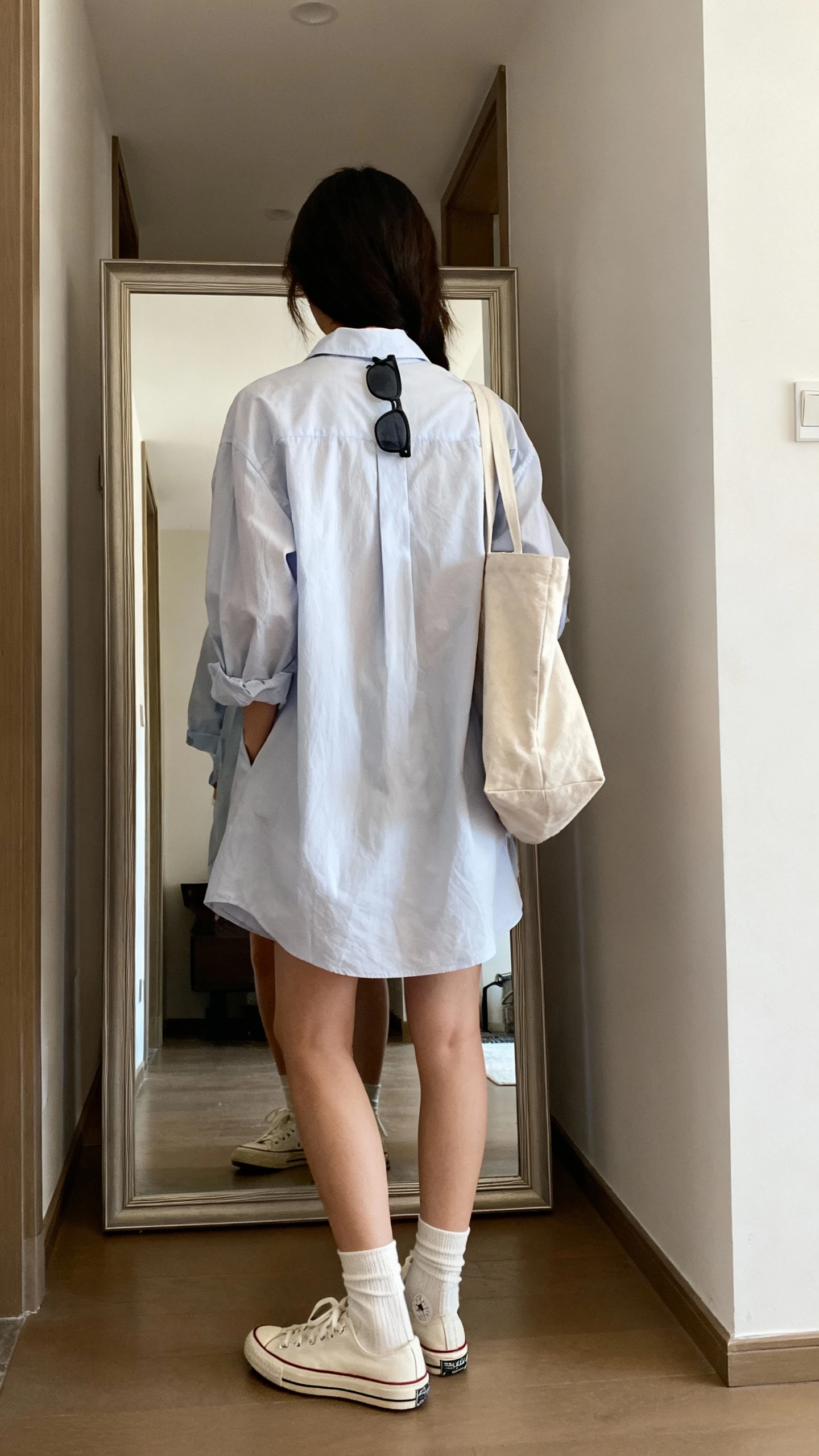 iPhone back view of a woman in a breezy shirt dress with classic low-top sneakers, ankle socks, a canvas tote, and simple sunglasses hooked at the collar; face not visible, hallway full-length mirror, natural daylight, iPhone photo quality.