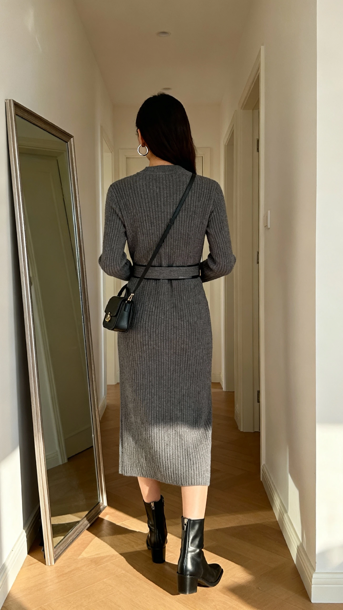 iPhone back view of a woman in a smart knit midi dress with a waist belt, sleek ankle boots, small crossbody bag, and subtle hoop earrings, face not visible, hallway full-length mirror, morning indoor lighting, casual iPhone photo.