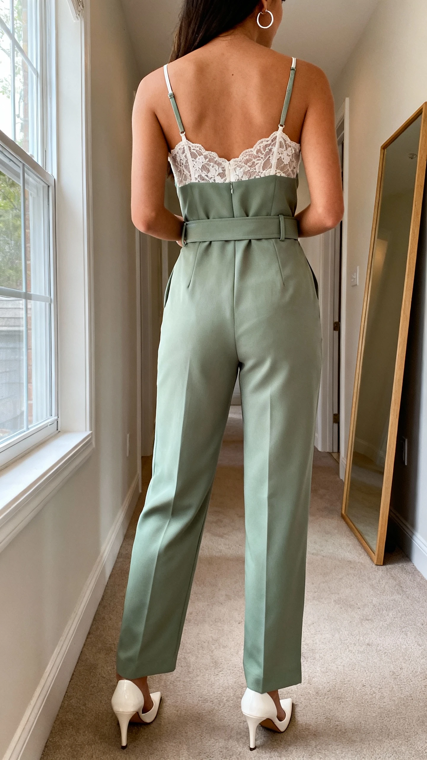 iPhone back view of a woman in a structured power jumpsuit with a romantic lace camisole peek, waist belt, pointed heels, and minimal hoop earrings, face not visible, hallway full-length mirror, natural daylight from window, iPhone photo quality.