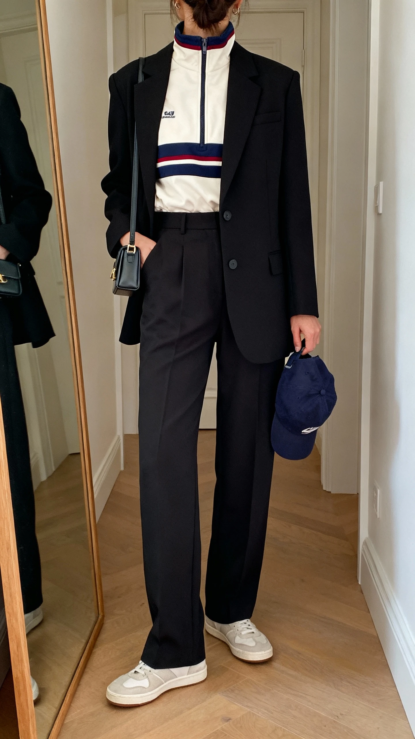 iPhone back view of a woman wearing a 90s-inspired track jacket layered under a tailored black blazer, high-waisted trousers, minimalist sneakers, baseball cap tucked in hand, and mini shoulder bag, face not visible, hallway full-length mirror, indoor lighting, iPhone photo quality.