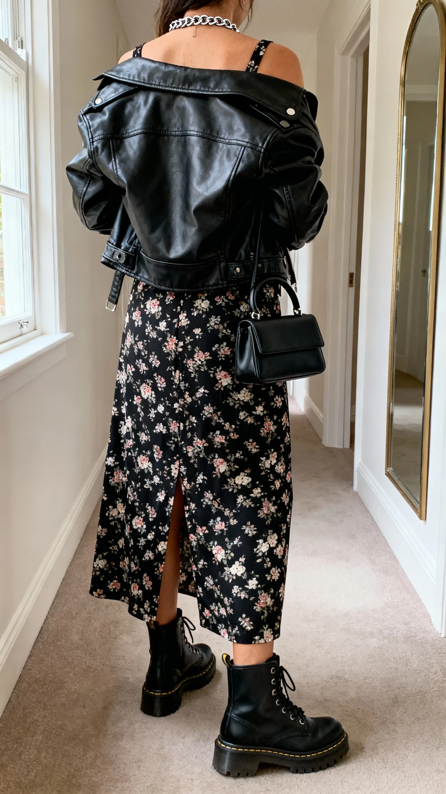 iPhone back view of a woman wearing a black-ground floral midi dress with a slit, leather moto jacket tossed over shoulders, chunky combat boots, silver chain necklace, small black top-handle bag, face not visible, hallway mirror, natural daylight from window, iPhone photo quality.