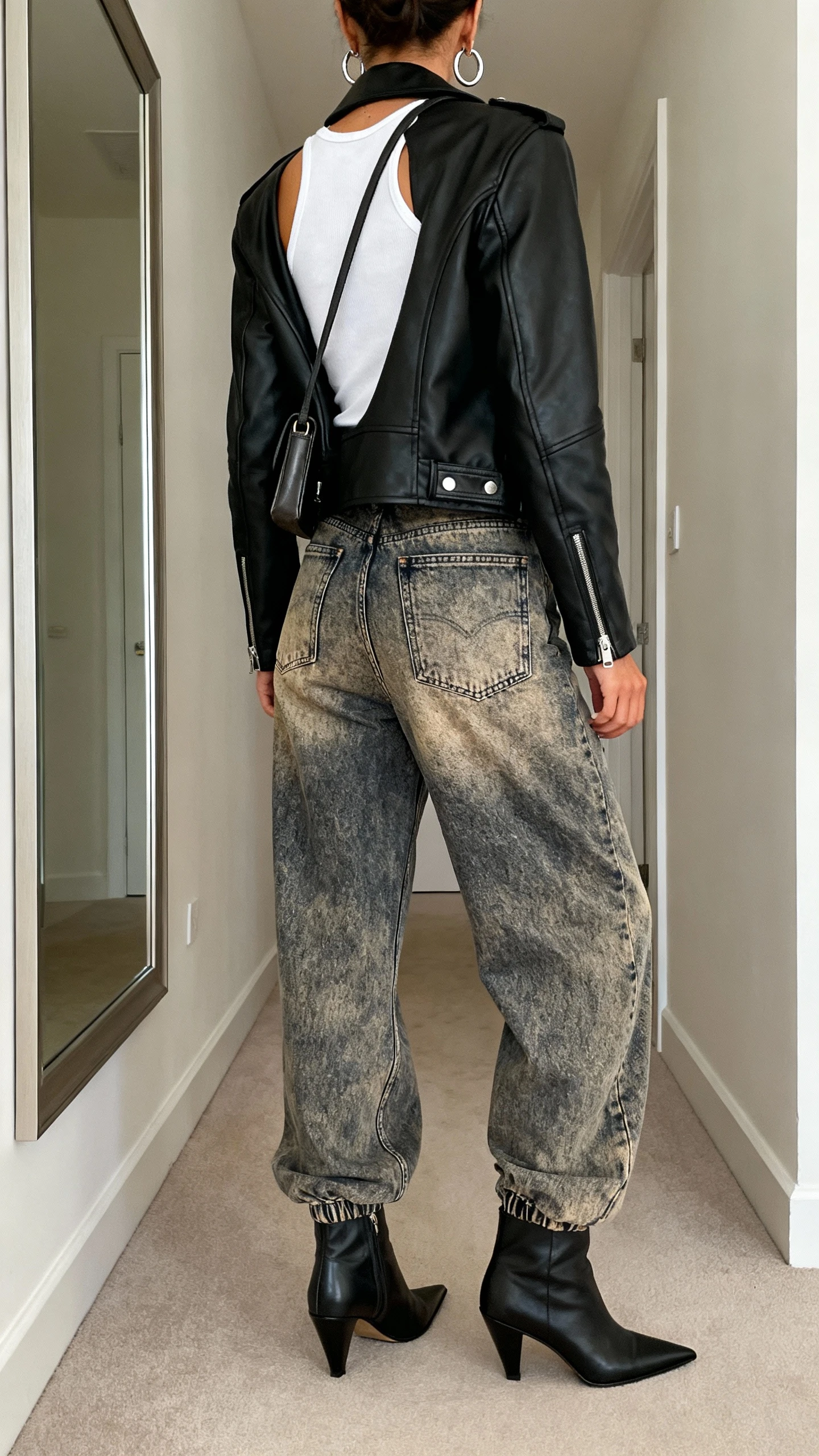 iPhone back view of a woman wearing a black leather moto jacket over a white tank, vintage-wash baggy denim, pointed ankle boots, silver hoop earrings, and a small shoulder bag, face not visible, hallway mirror, natural daylight, casual iPhone photo.