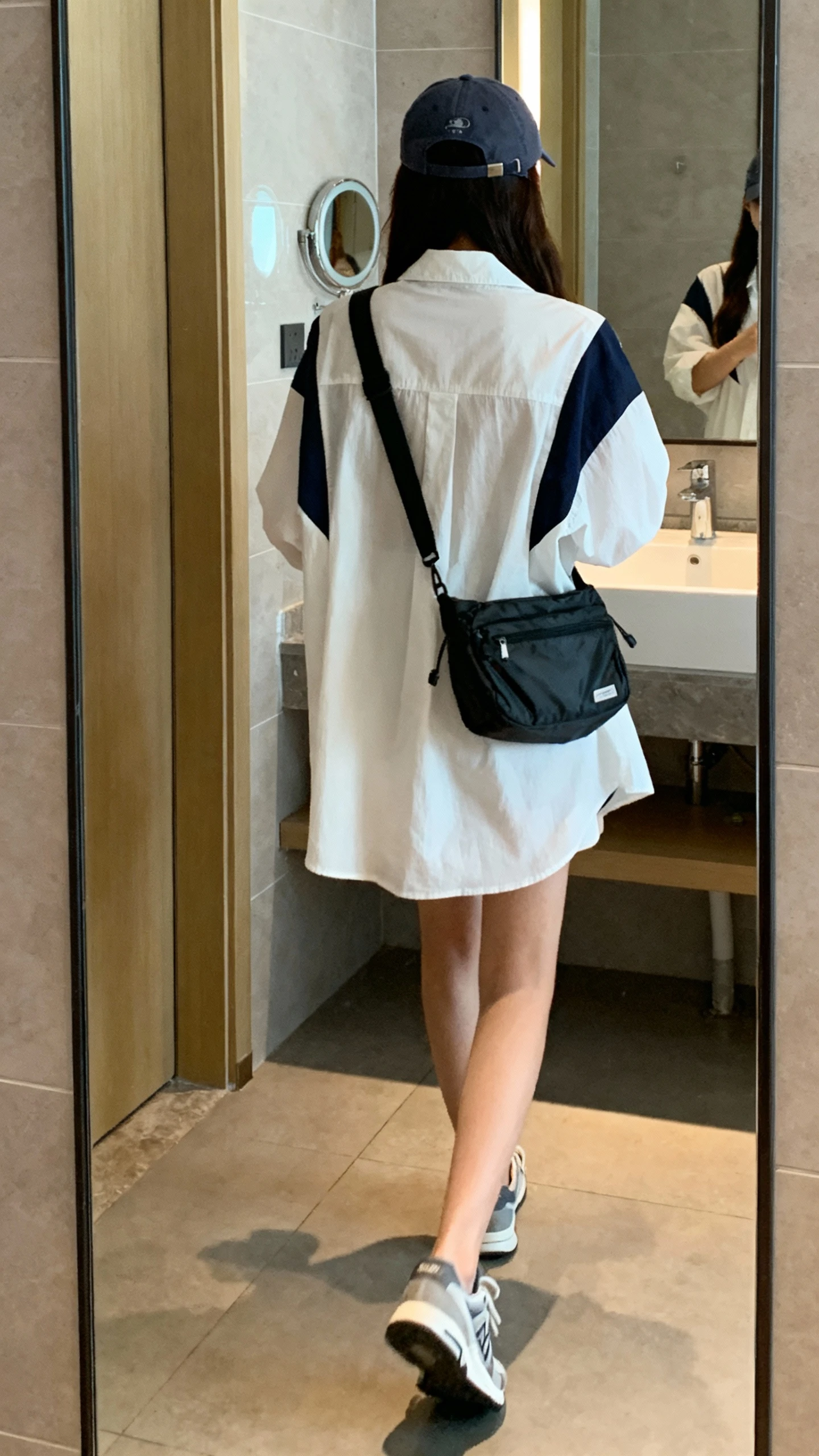 iPhone back view of a woman wearing a breezy shirtdress with sporty accents: baseball cap, crossbody nylon bag, low-profile sneakers, face not visible, walking pose in bathroom mirror with soft natural light, casual iPhone photo.