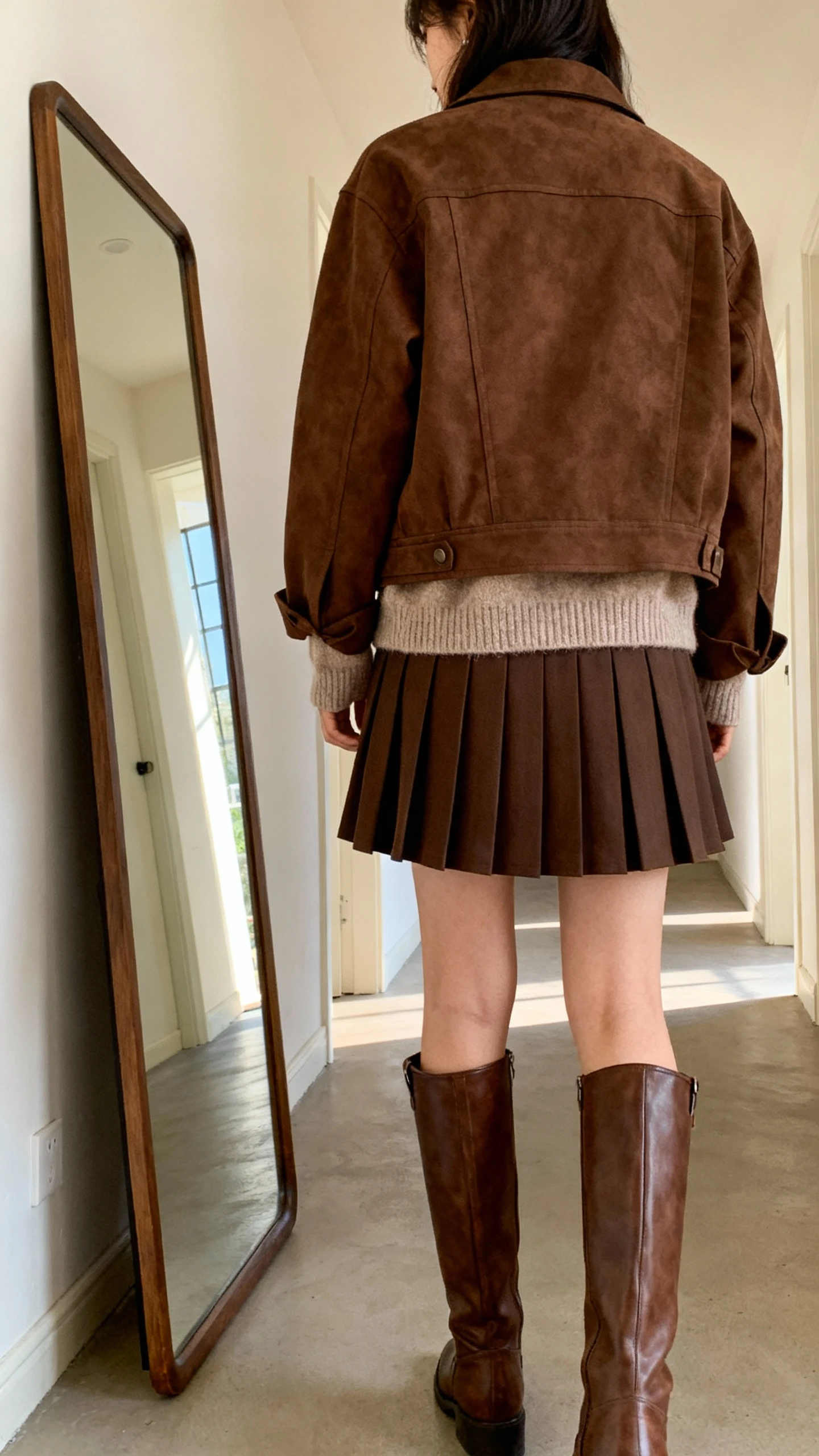 iPhone back view of a woman wearing a brown jacket with a pleated skirt, a soft cardigan layered under the jacket, and knee-high brown boots, face not visible, hallway full-length mirror, natural daylight, iPhone photo quality.