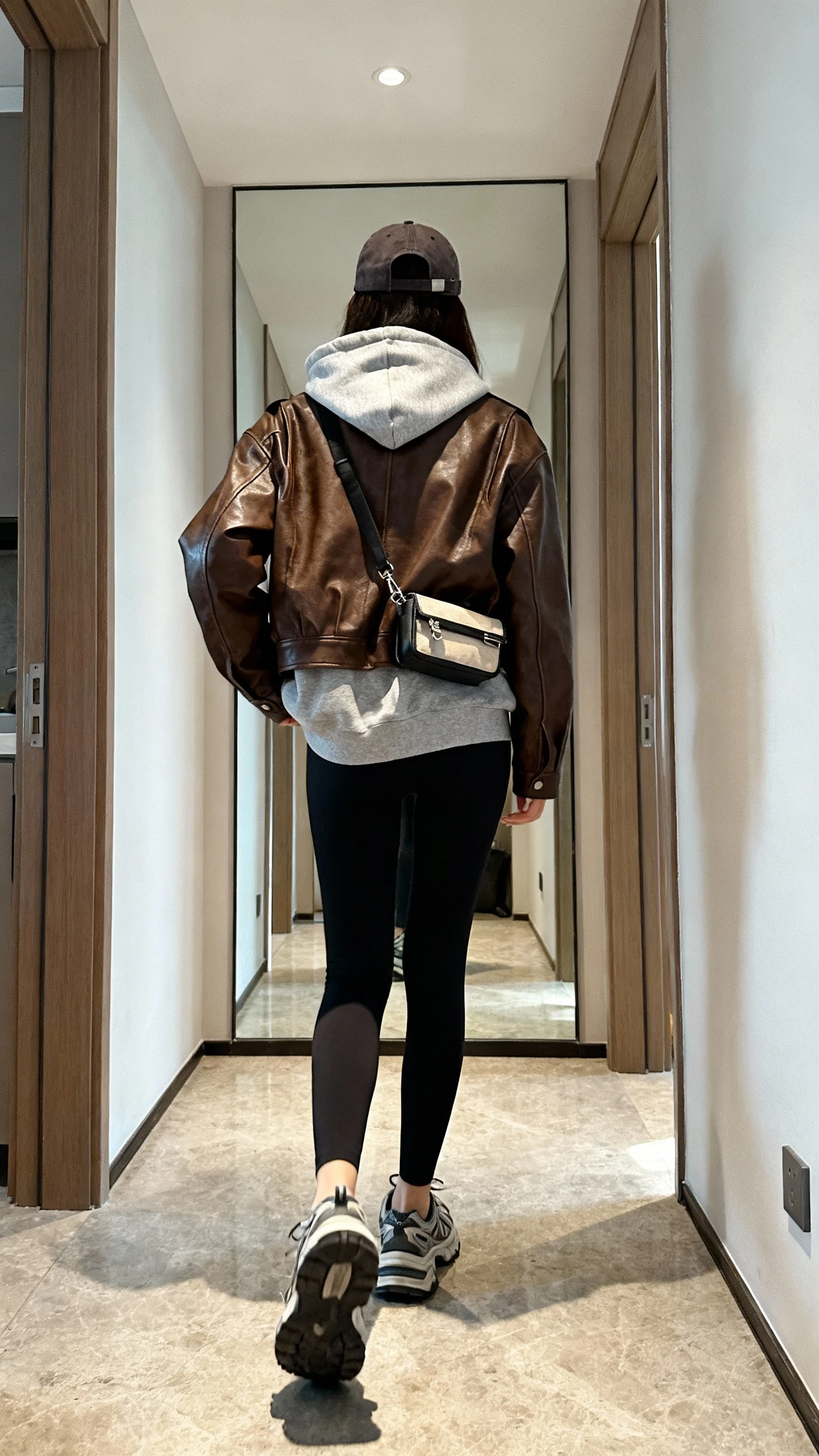 iPhone back view of a woman wearing a brown leather jacket with a grey hoodie layered underneath, black leggings, chunky dad sneakers, baseball cap, and a small belt bag, face not visible, walking pose in hallway mirror, indoor daylight, iPhone photo quality.