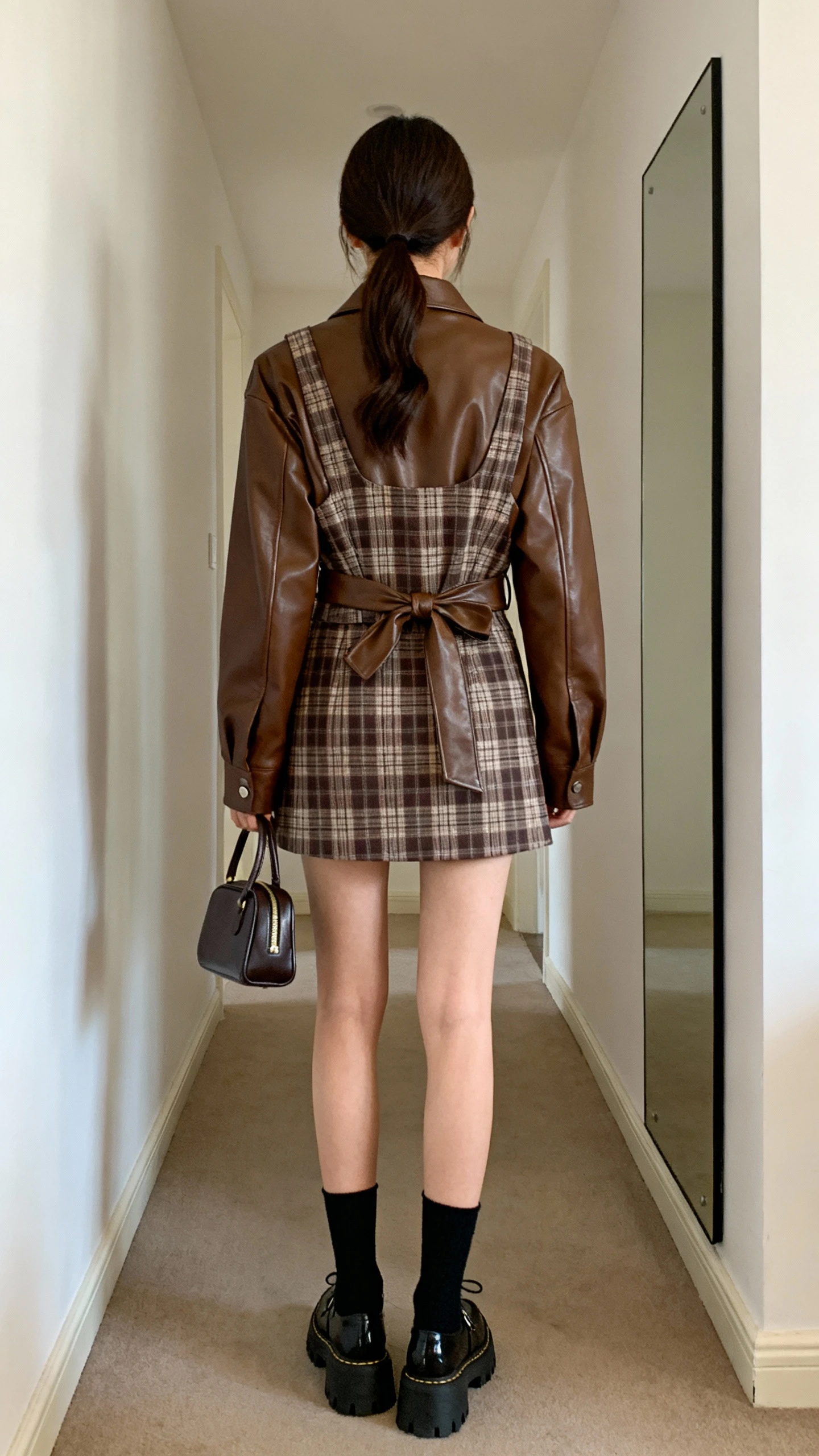 iPhone back view of a woman wearing a brown leather jacket over a plaid mini skirt set (matching jacket tied at waist or vest), black crew socks with chunky Mary Janes, a structured mini handbag, and hair in a low pony, face not visible, hallway full-length mirror, morning light, iPhone photo quality.