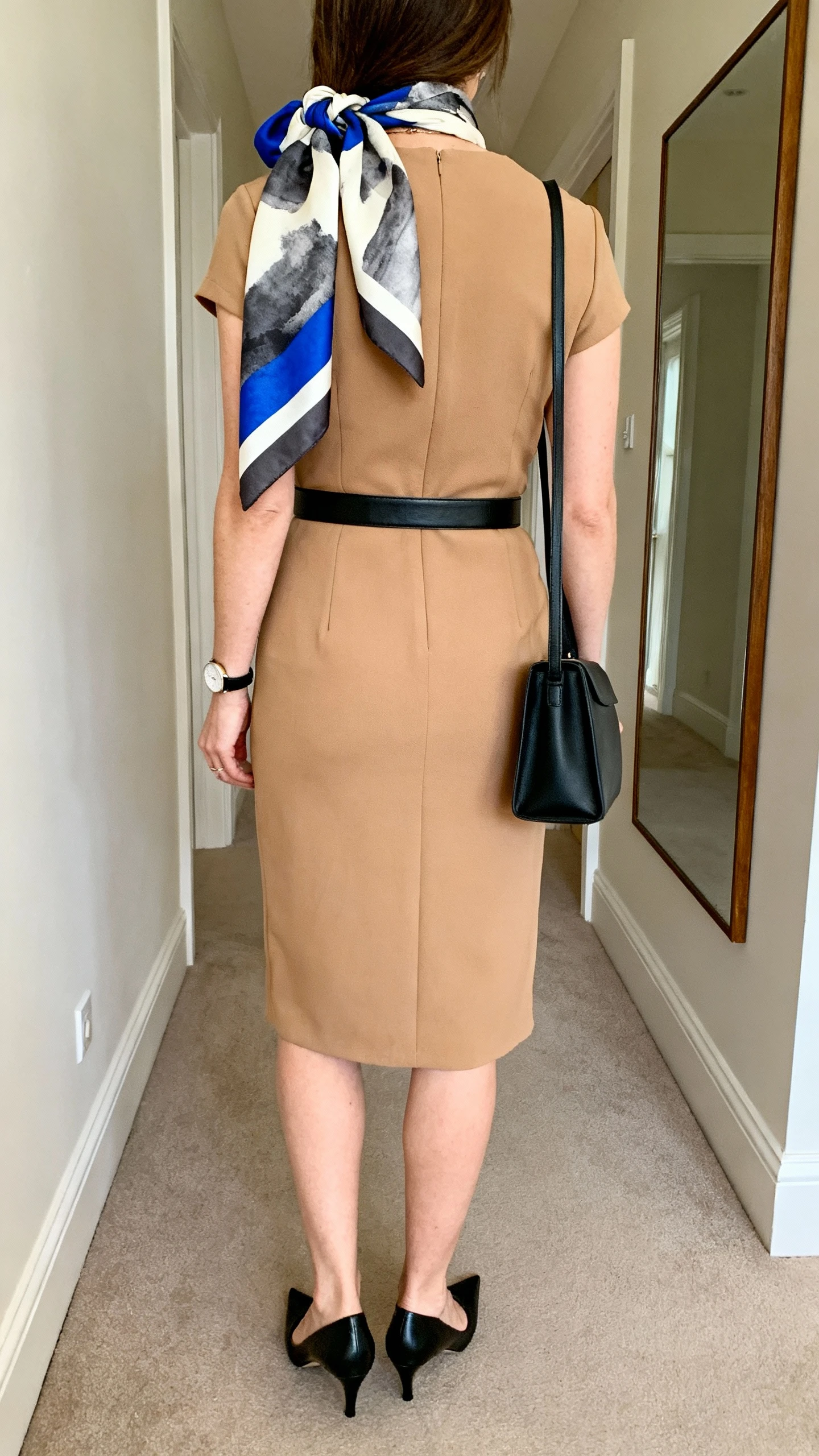 iPhone back view of a woman wearing a camel sheath dress with a black minimalist leather belt, black pointed heels, a gallery-worthy abstract-print silk scarf in cobalt, charcoal, and ivory knotted at the shoulder, slim black tote, and a simple watch, face not visible, hallway mirror, soft indoor daylight, iPhone photo quality.