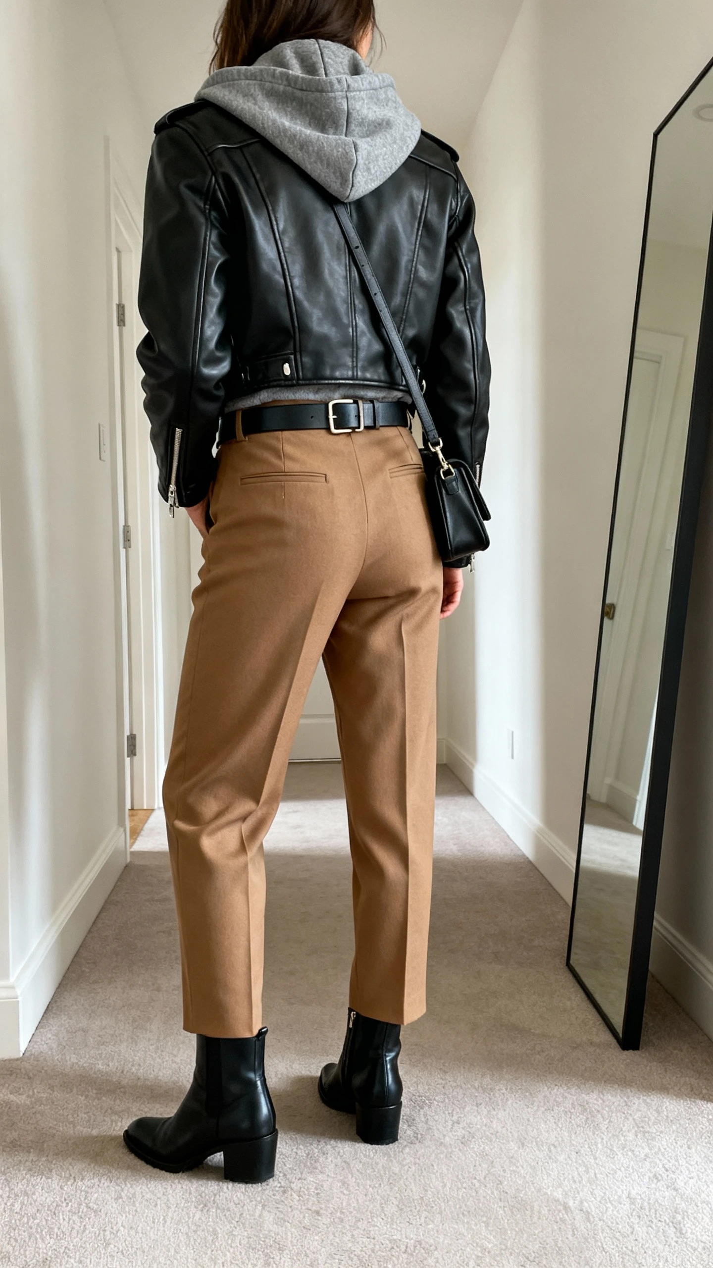 iPhone back view of a woman wearing a charcoal hoodie layered with a black leather biker jacket, tailored camel trousers, black ankle boots, belt, crossbody bag, face not visible, hallway full-length mirror, indoor daylight, iPhone photo quality.
