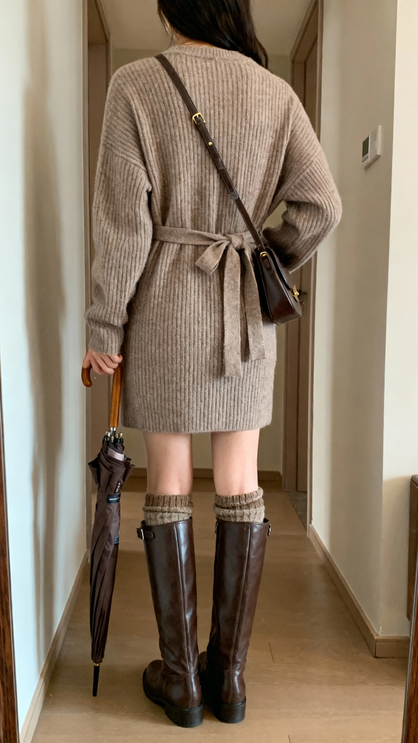 iPhone back view of a woman wearing a cozy ribbed sweater dress with a waist tie, tall knee-high leather boots, wool socks peeking, crossbody bag, and a compact foldable umbrella tucked under arm, face not visible, hallway mirror with soft indoor lighting, iPhone photo quality.