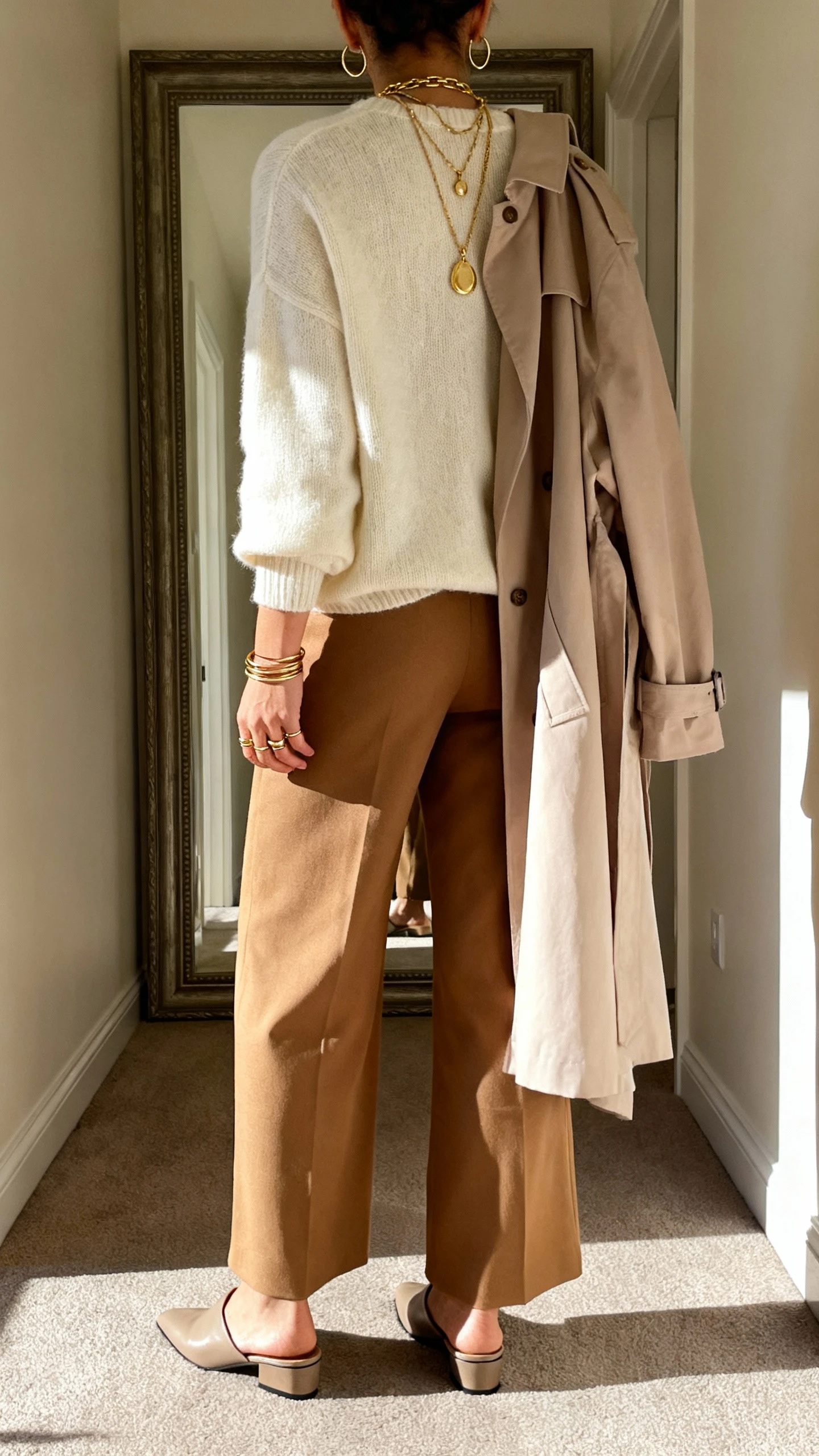 iPhone back view of a woman wearing a cream cashmere sweater, tan wide-leg trousers, soft beige trench draped over shoulders, sunlit gold jewelry (layered pendant necklaces, slim stacking rings, thin hoops), and neutral leather mules, face not visible, full-length hallway mirror, warm indoor light, iPhone photo quality