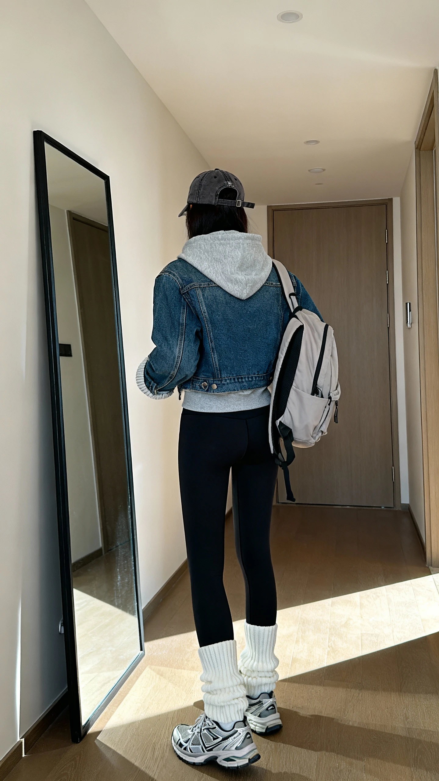 iPhone back view of a woman wearing a denim jacket with black high-waisted leggings, gray hoodie peeking at the hood and cuffs, chunky white socks and sporty trainers, baseball cap and simple backpack, face not visible, hallway full-length mirror, morning light, iPhone photo quality