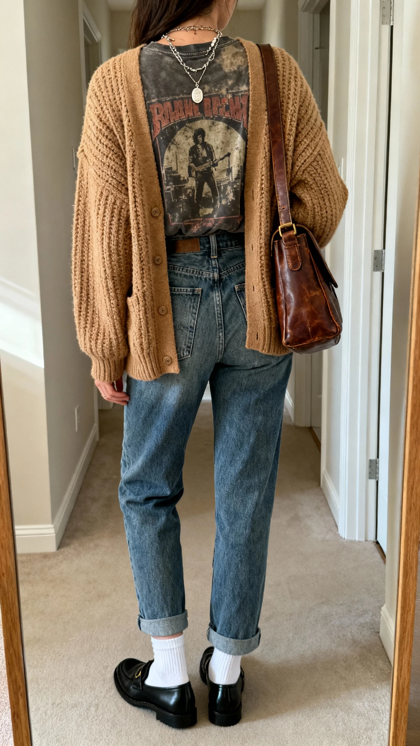 iPhone back view of a woman wearing a faded vintage band tee tucked into high-waisted mom jeans, chunky knit camel cardigan, black loafers with white socks, layered pendant necklaces, and a thrifted leather shoulder bag, face not visible, hallway full-length mirror, soft morning light, iPhone photo quality.