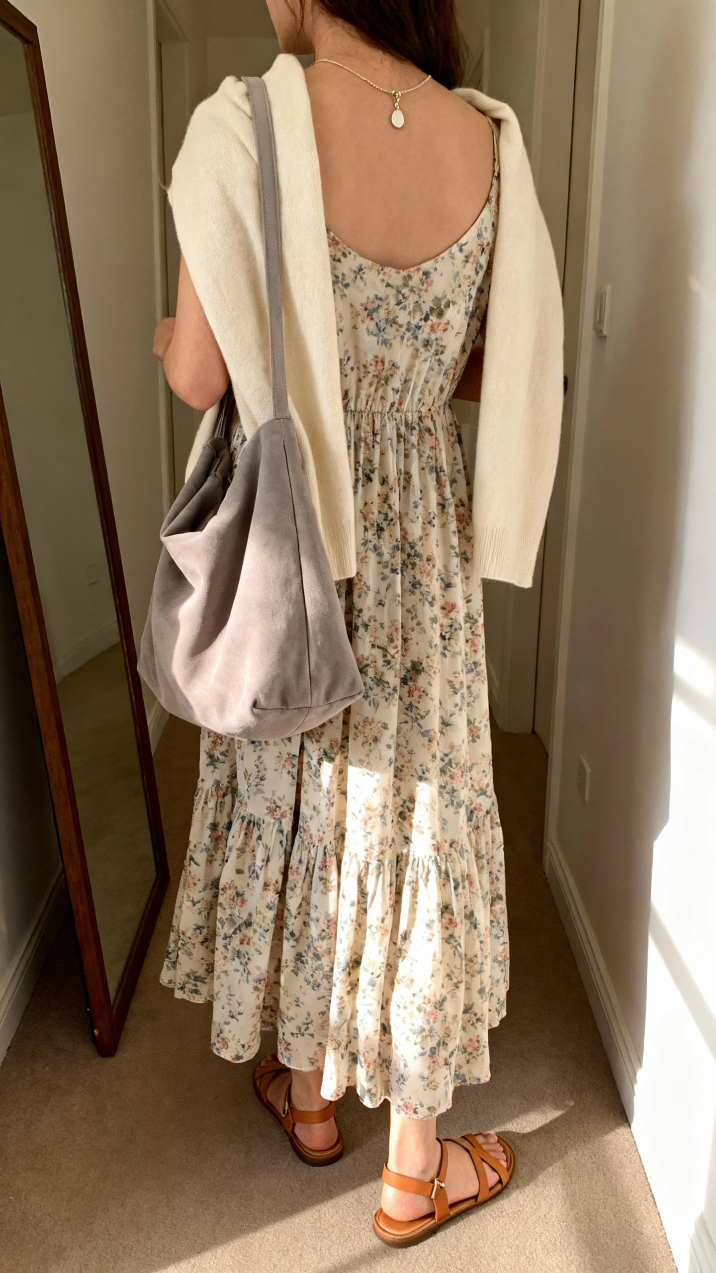 iPhone back view of a woman wearing a flowy floral maxi dress, soft cream cardigan draped over shoulders, delicate pendant necklace, strappy tan sandals, carrying a slouchy dove-gray soft shoulder bag, face not visible, hallway full-length mirror, warm indoor lighting, iPhone photo quality.