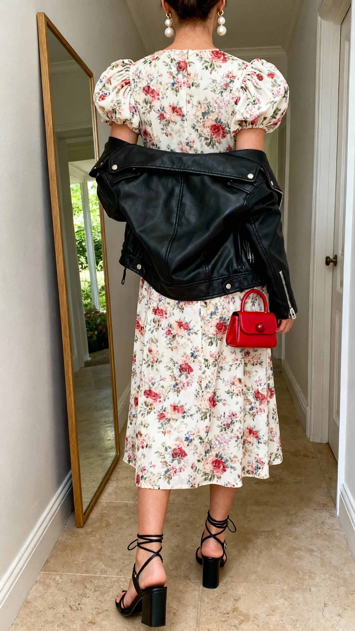 iPhone back view of a woman wearing a garden party romantic look with a bite: floral puff-sleeve midi dress, black leather moto jacket over shoulders, strappy black block-heel sandals, pearl drop earrings, and a tiny cherry-red top-handle bag, face not visible, hallway full-length mirror, afternoon natural light, iPhone photo quality