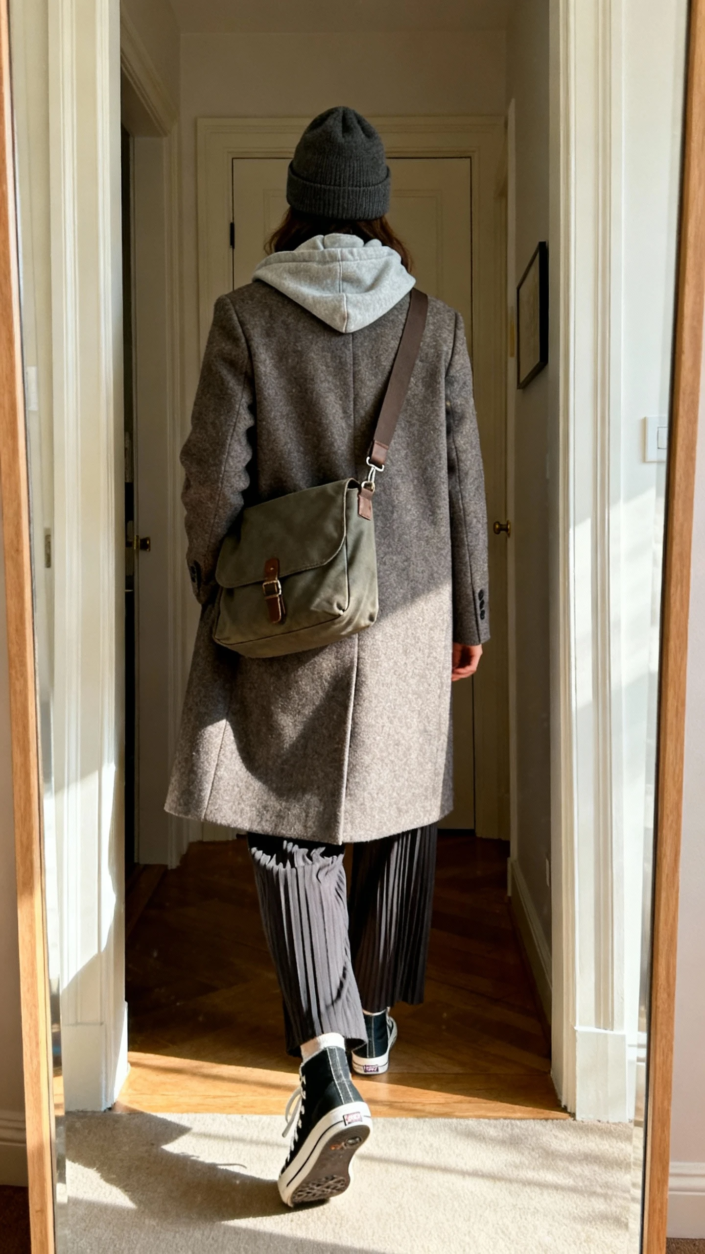 iPhone back view of a woman wearing a medium messenger bag crossbody, tailored wool coat over a hoodie, pleated trousers, skate-style sneakers, and a beanie; face not visible, walking pose in entryway mirror, afternoon indoor light, iPhone photo quality.