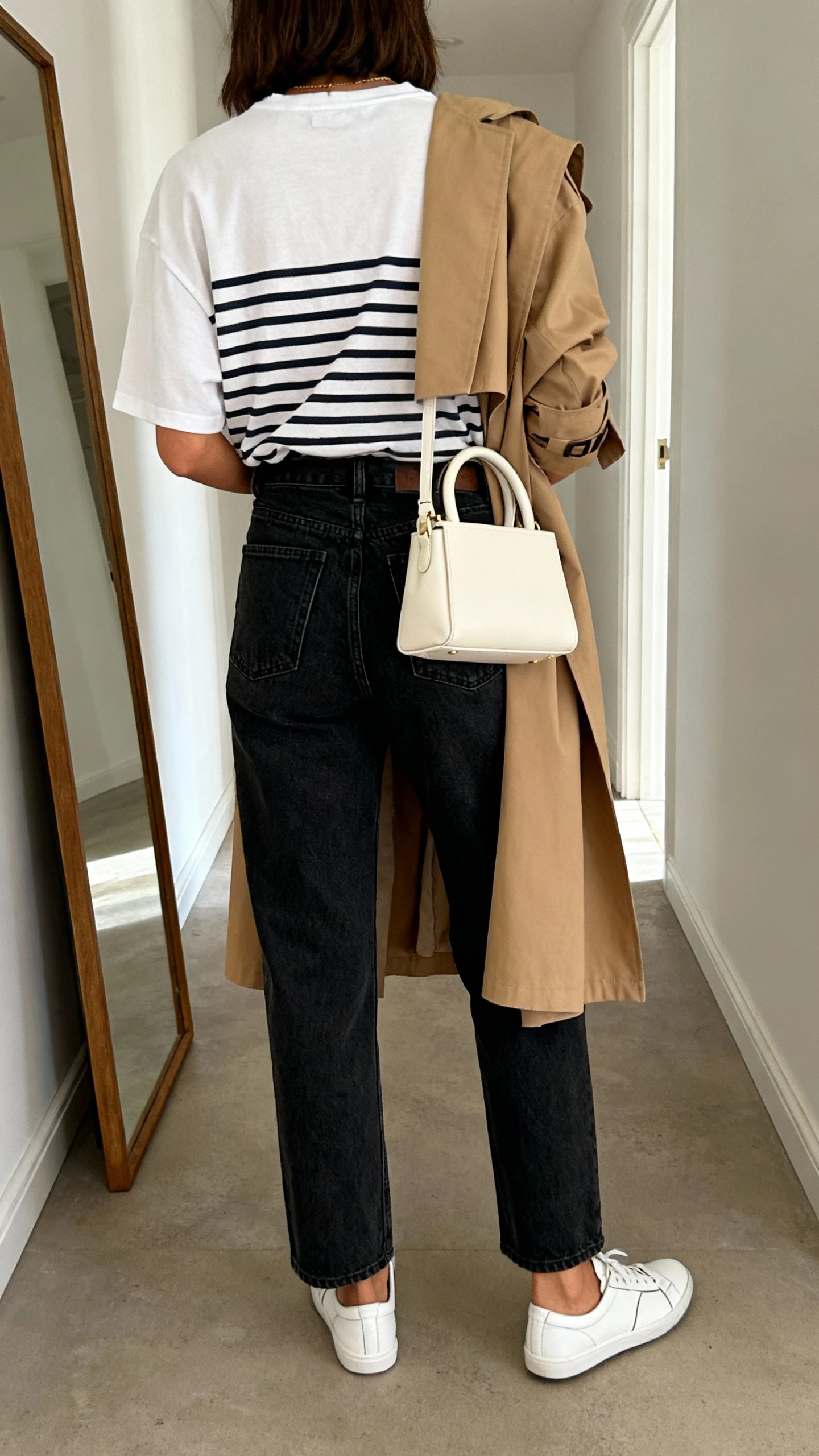 iPhone back view of a woman wearing a mini top-handle bag in cream, boxy striped Breton tee tucked into high-waist dark-wash straight jeans, lightweight trench in camel draped over shoulders, and white leather sneakers; face not visible, hallway full-length mirror, indoor daylight, iPhone photo quality.