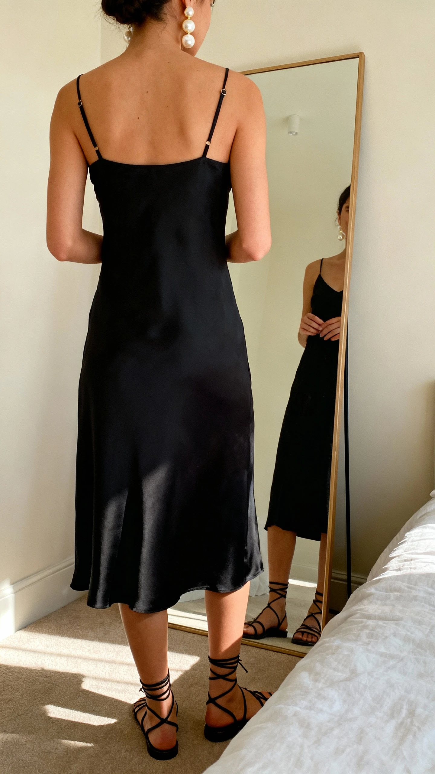 iPhone back view of a woman wearing a minimal black slip dress with delicate straps, strappy sandals, and oversized pearl statement earrings that graze the collarbone, face not visible, full-length mirror in a softly lit bedroom, natural afternoon light, iPhone photo quality.