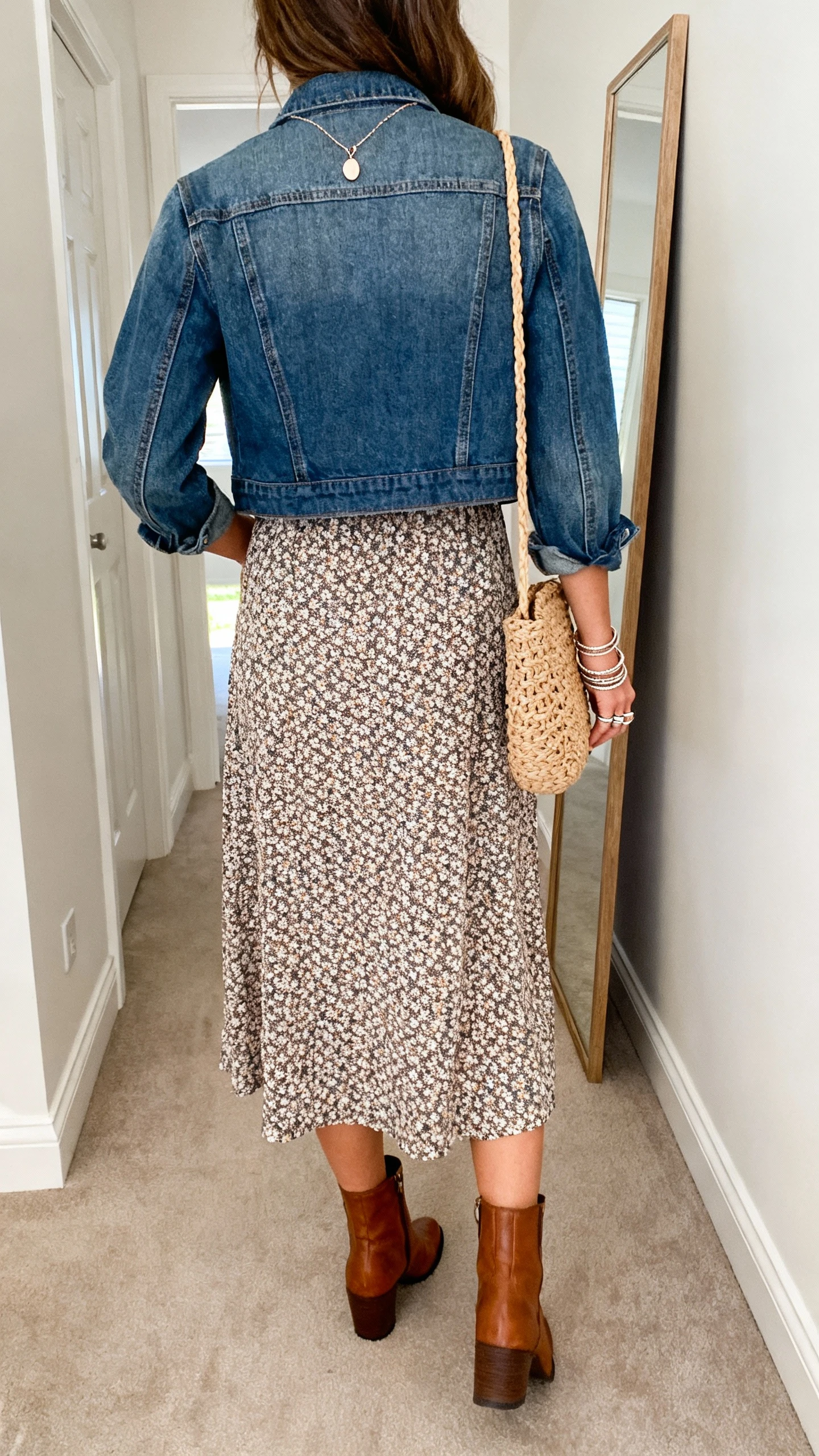 iPhone back view of a woman wearing a printed midi dress with leather ankle boots, a cropped denim jacket, a delicate pendant necklace, stackable rings, and a woven shoulder bag; face not visible, hallway full-length mirror, natural daylight, iPhone photo quality.