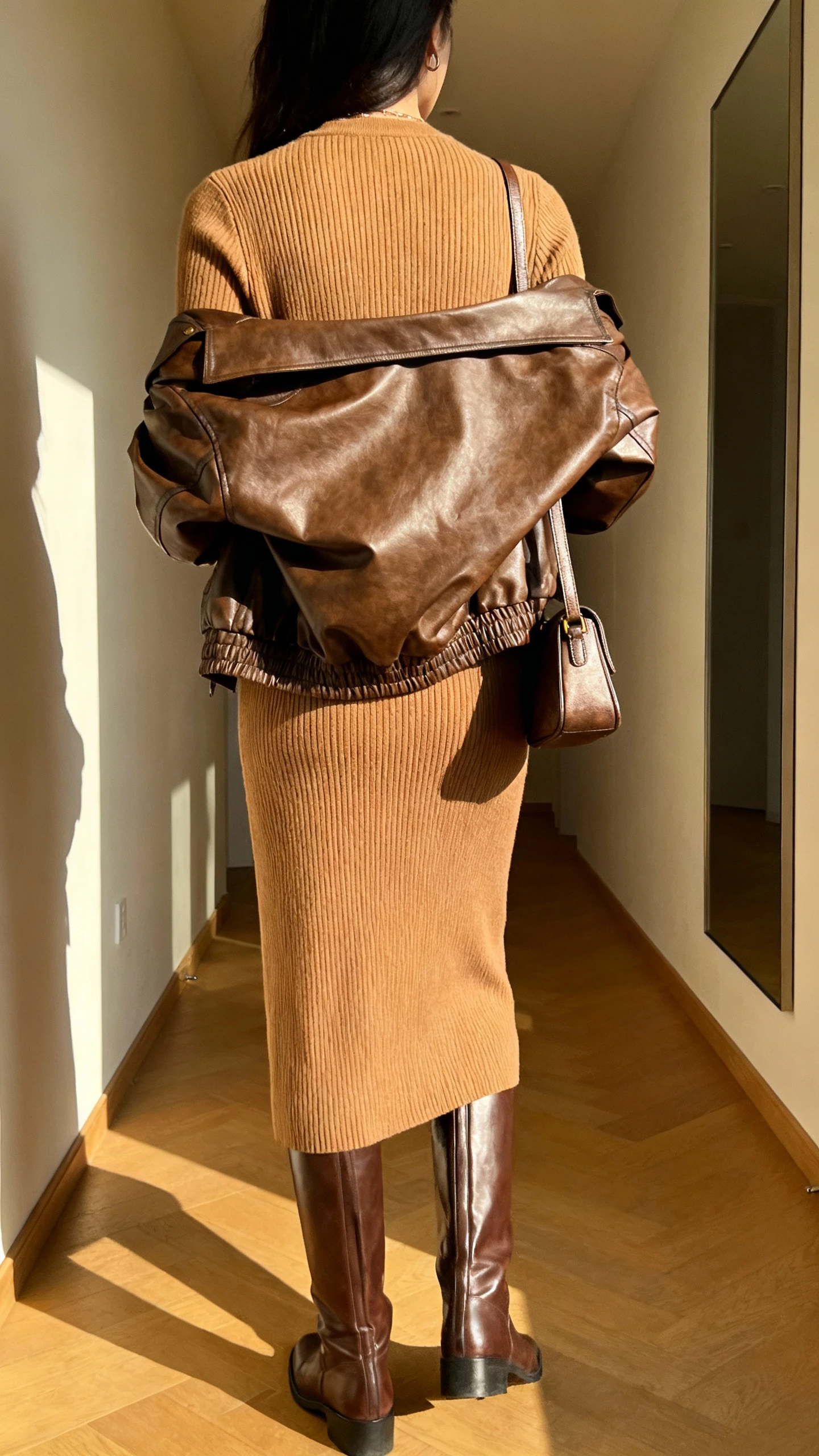 iPhone back view of a woman wearing a ribbed knit midi dress in tan with a brown leather jacket dupe draped over shoulders, knee-high leather boots, and a small shoulder bag, face not visible, hallway mirror, warm afternoon light, iPhone photo quality