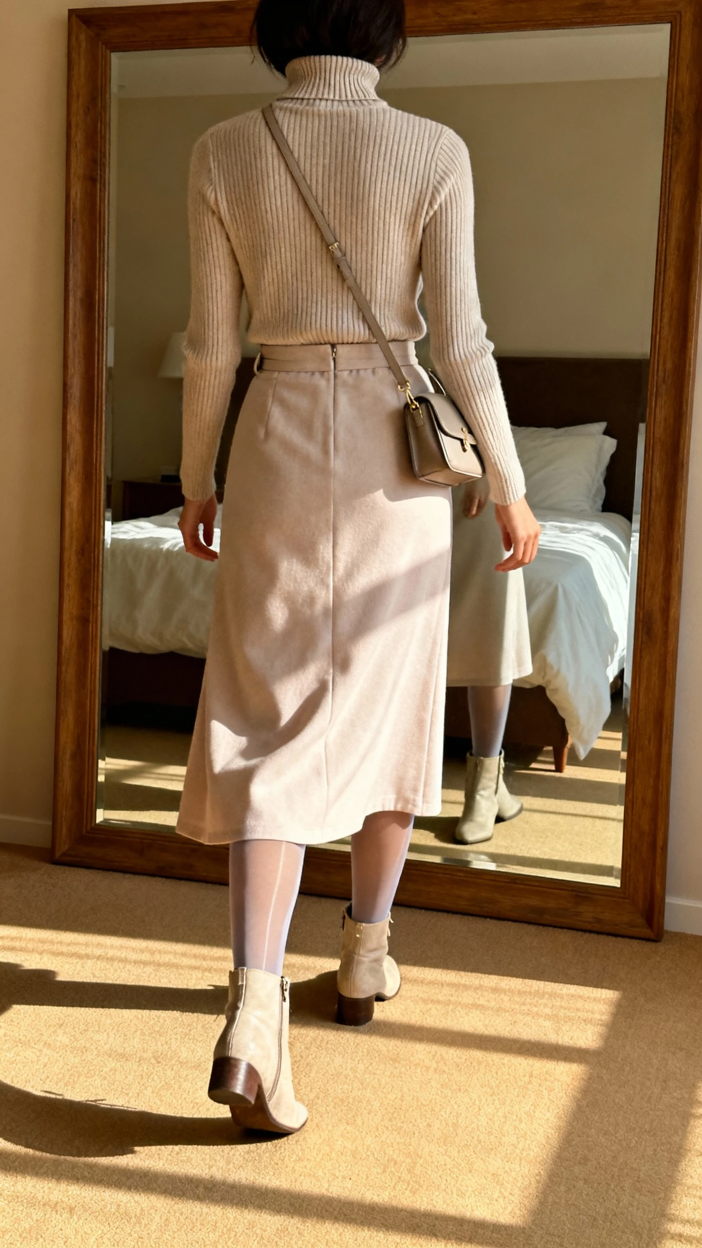 iPhone back view of a woman wearing a ribbed turtleneck tucked into a soft midi skirt with sheer tights, refined ankle boots, and a small shoulder bag, face not visible, walking pose, bedroom mirror, gentle afternoon light, iPhone photo quality.