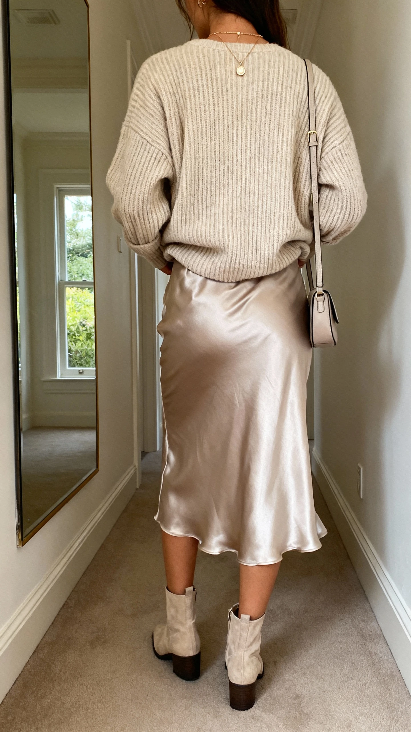 iPhone back view of a woman wearing a satin slip skirt with a chunky ribbed knit sweater half-tucked, ankle boots, delicate pendant necklace, small shoulder bag, face not visible, hallway mirror, natural daylight from window, iPhone photo quality
