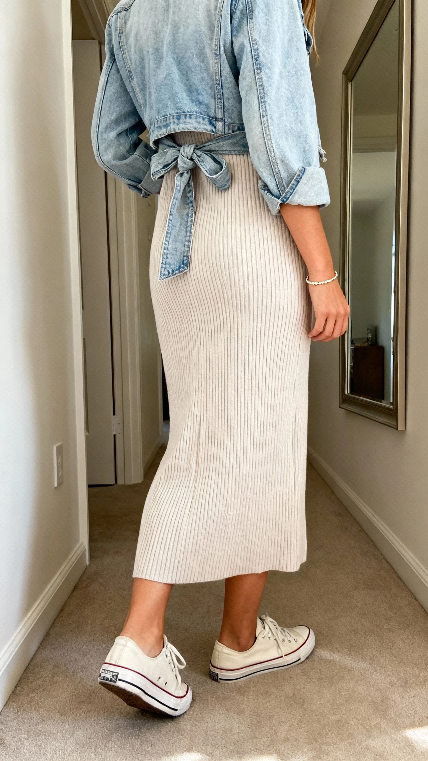 iPhone back view of a woman wearing a soft ribbed knit midi dress with classic low-top sneakers, lightweight denim jacket tied at the waist, delicate bracelet, face not visible, hallway mirror, indoor daylight, iPhone photo quality.