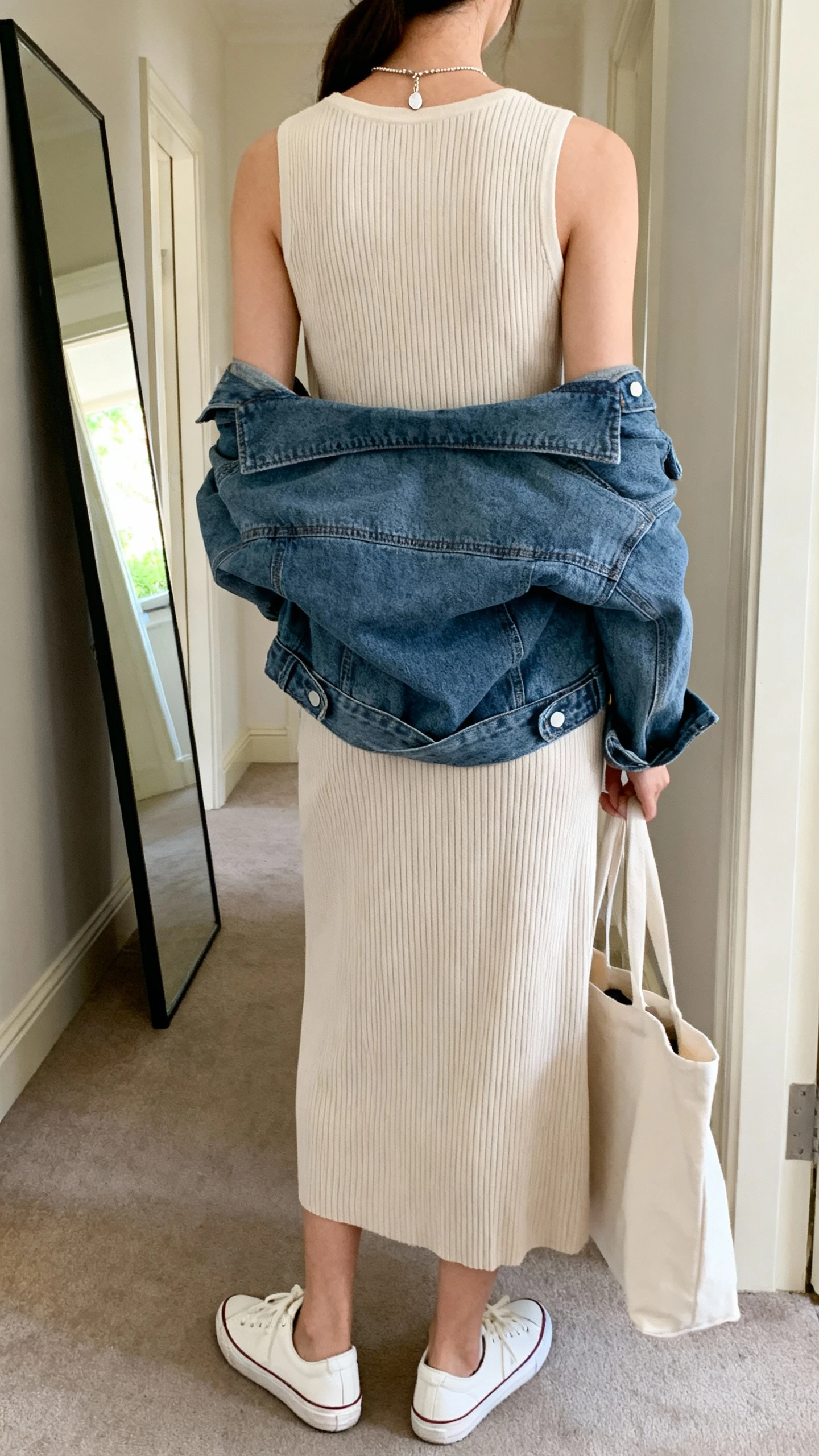 iPhone back view of a woman wearing a soft ribbed knit midi dress with a denim jacket tied at the waist, low-top white sneakers, simple pendant necklace, tote bag in hand, face not visible, hallway full-length mirror, natural daylight, iPhone photo quality