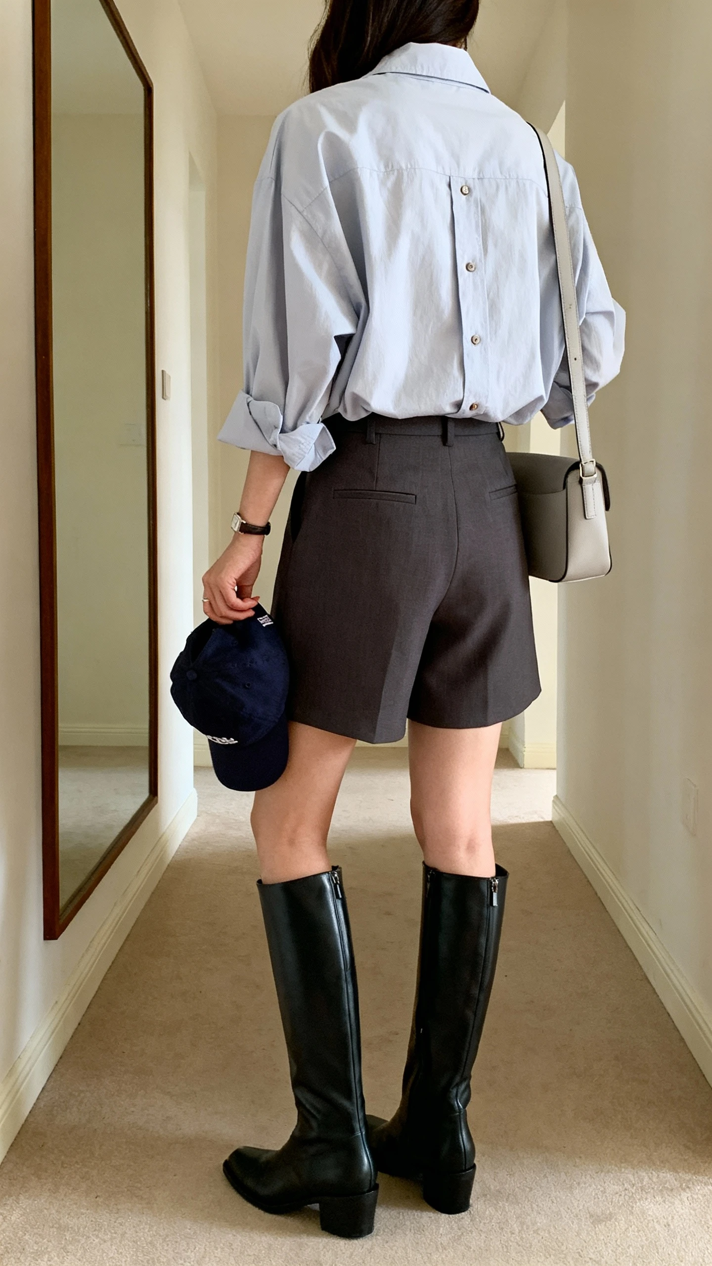 iPhone back view of a woman wearing an oversized button-down shirt with tailored shorts and over-the-knee boots, face not visible, hallway mirror, natural daylight, iPhone photo quality; baseball cap in hand and simple shoulder bag.